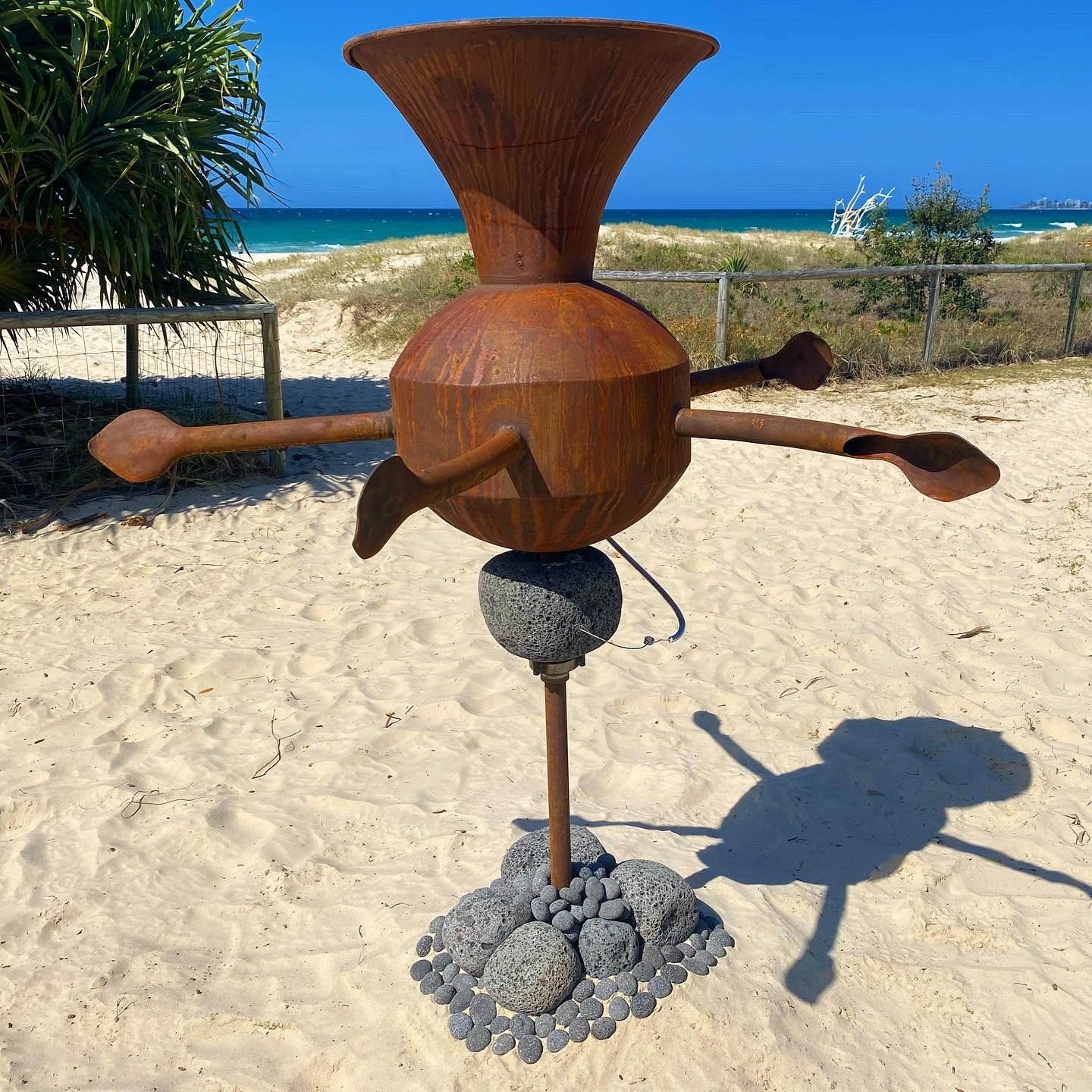 Rusty metal sculpture with propeller-like arms on sandy beach with ocean in background.