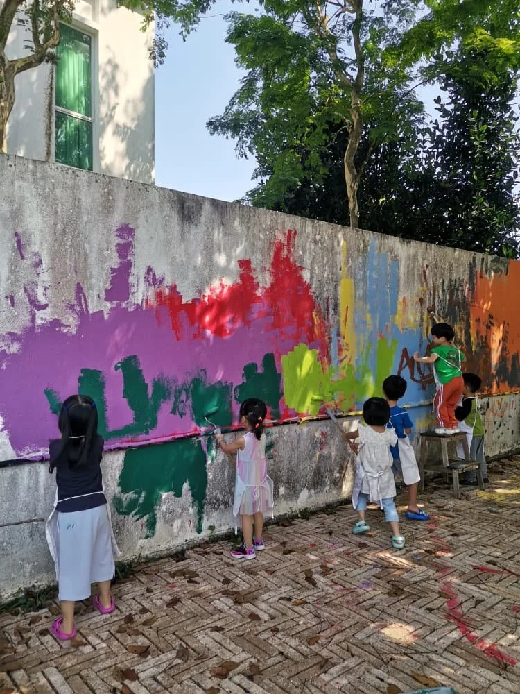 Children painting a colorful mural on a concrete wall outdoors, with trees and a building in the background.