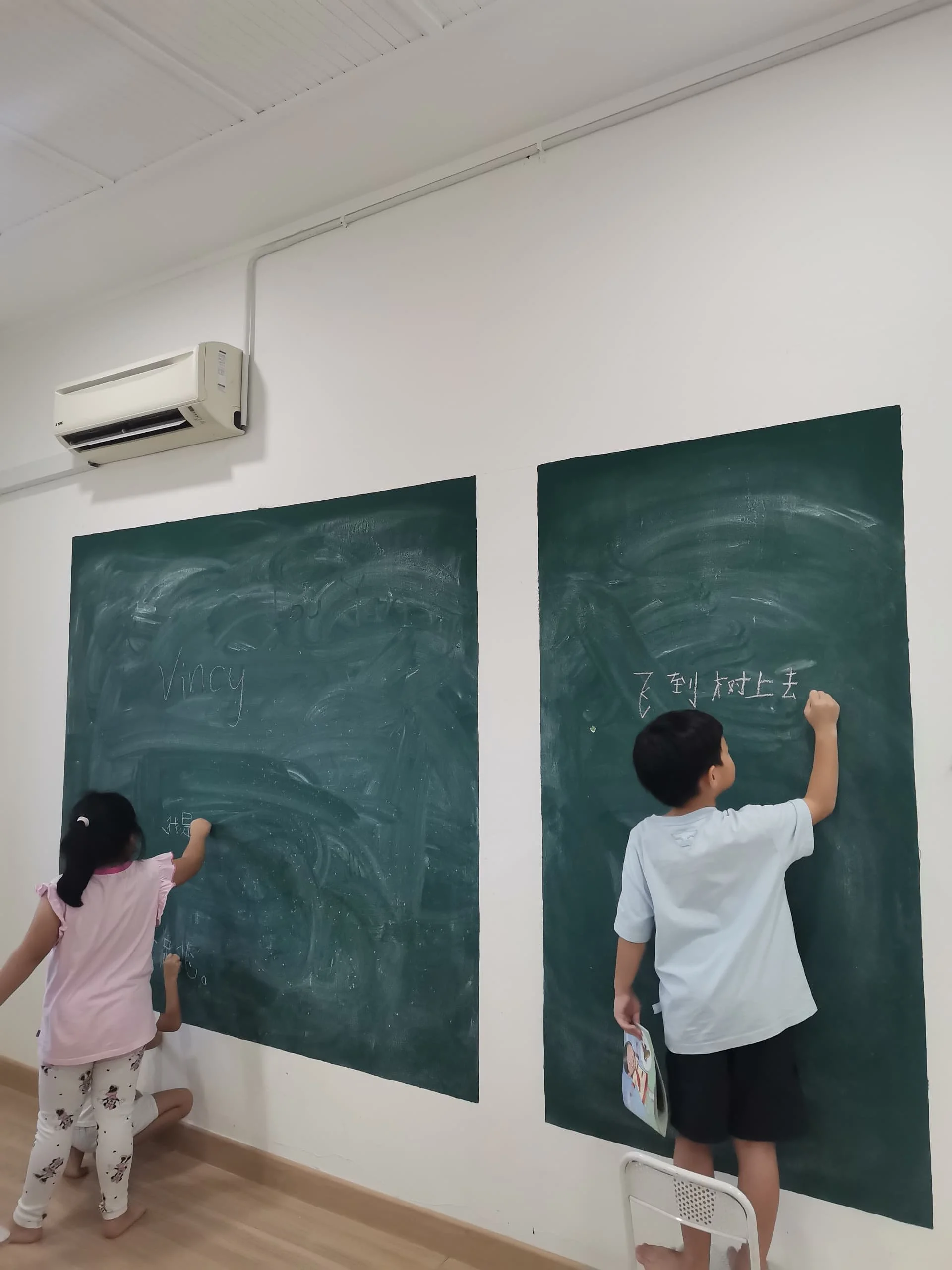 Two children writing on black chalkboards in a classroom, one girl wearing pink pajamas and a boy in a white shirt and dark shorts, with a girl dressed in black and white standing nearby.