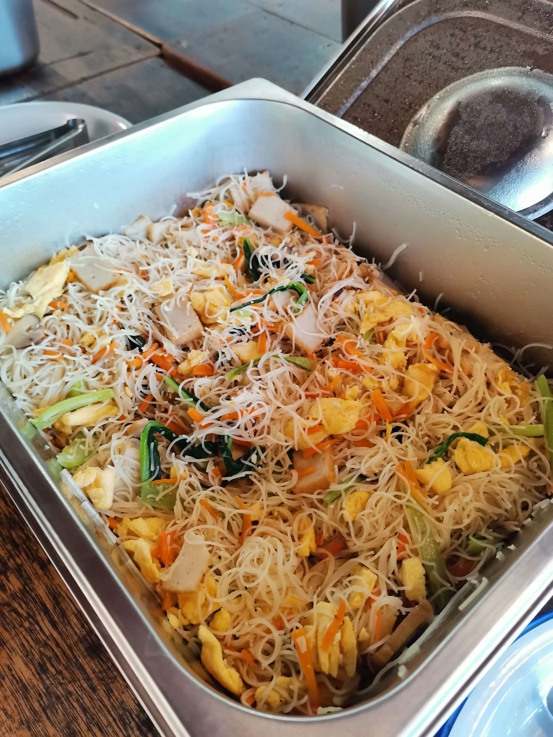Stainless steel container filled with cooked Japanese-style stir-fried noodles with vegetables, scrambled eggs, and pieces of chicken or tofu, on a wooden table.