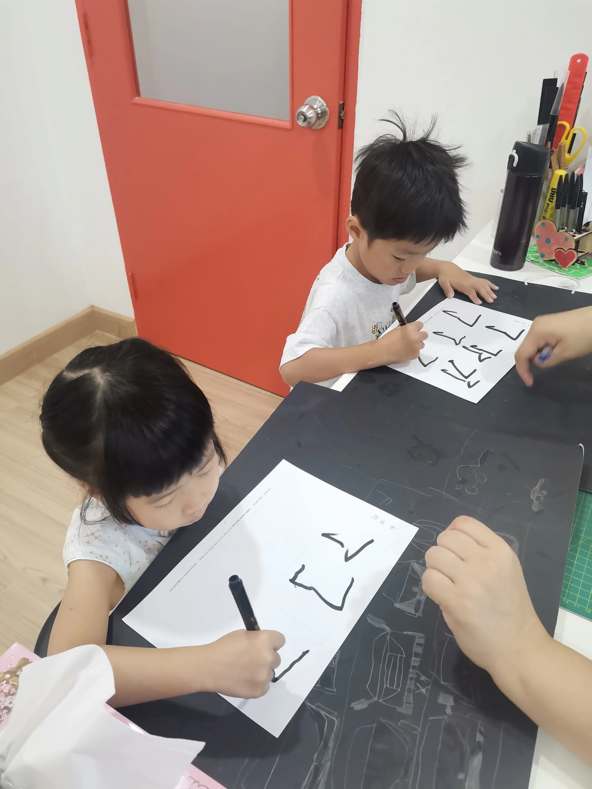 Two young children sitting at a table, practicing writing the letter 'L' on sheets of paper with black markers. The scene appears to be in a classroom or art space, with a red door and various writing supplies on a nearby counter.