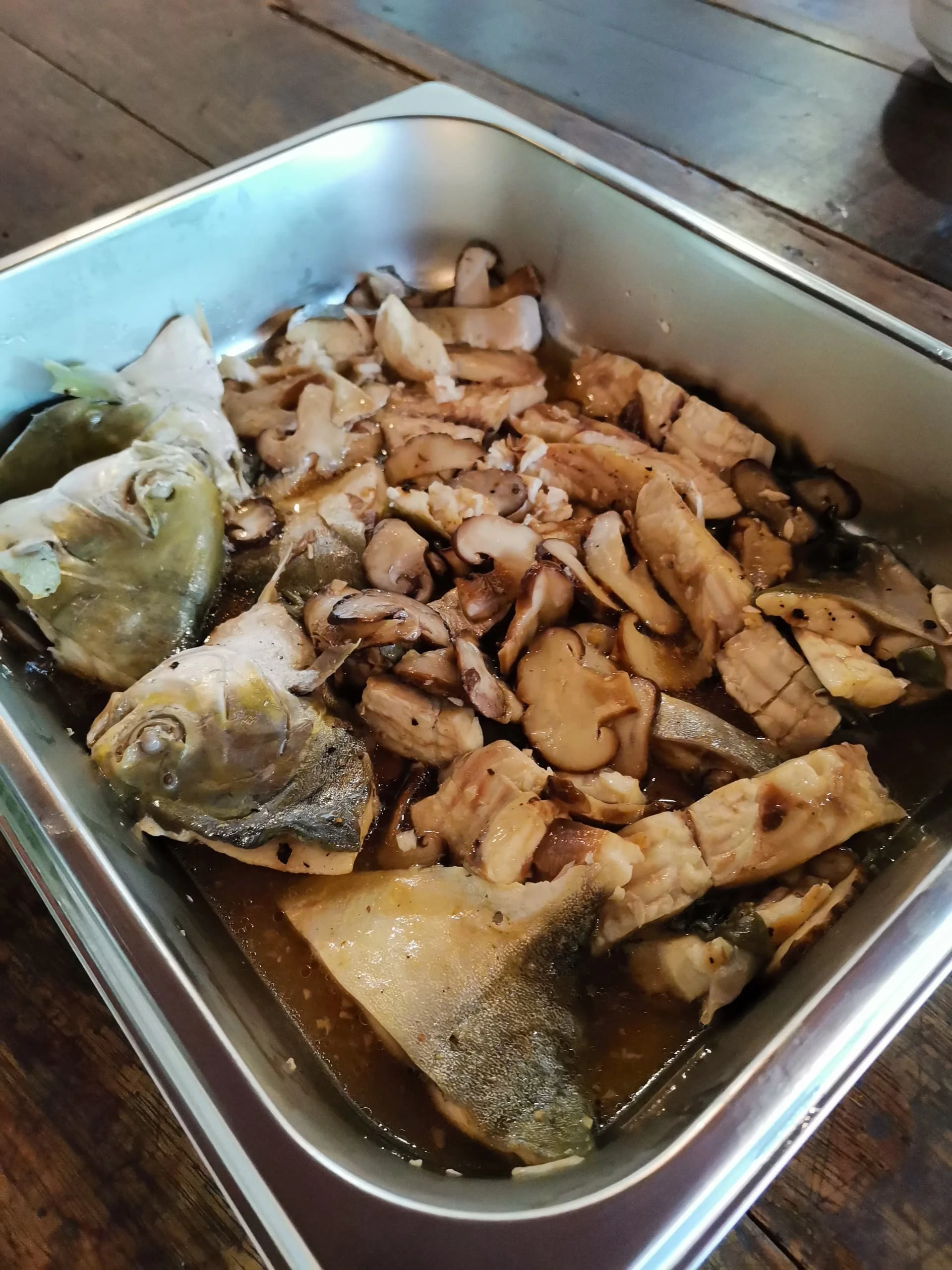 A metal container with cooked fish, mushrooms, and chopped vegetables inside, resting on a wooden surface.
