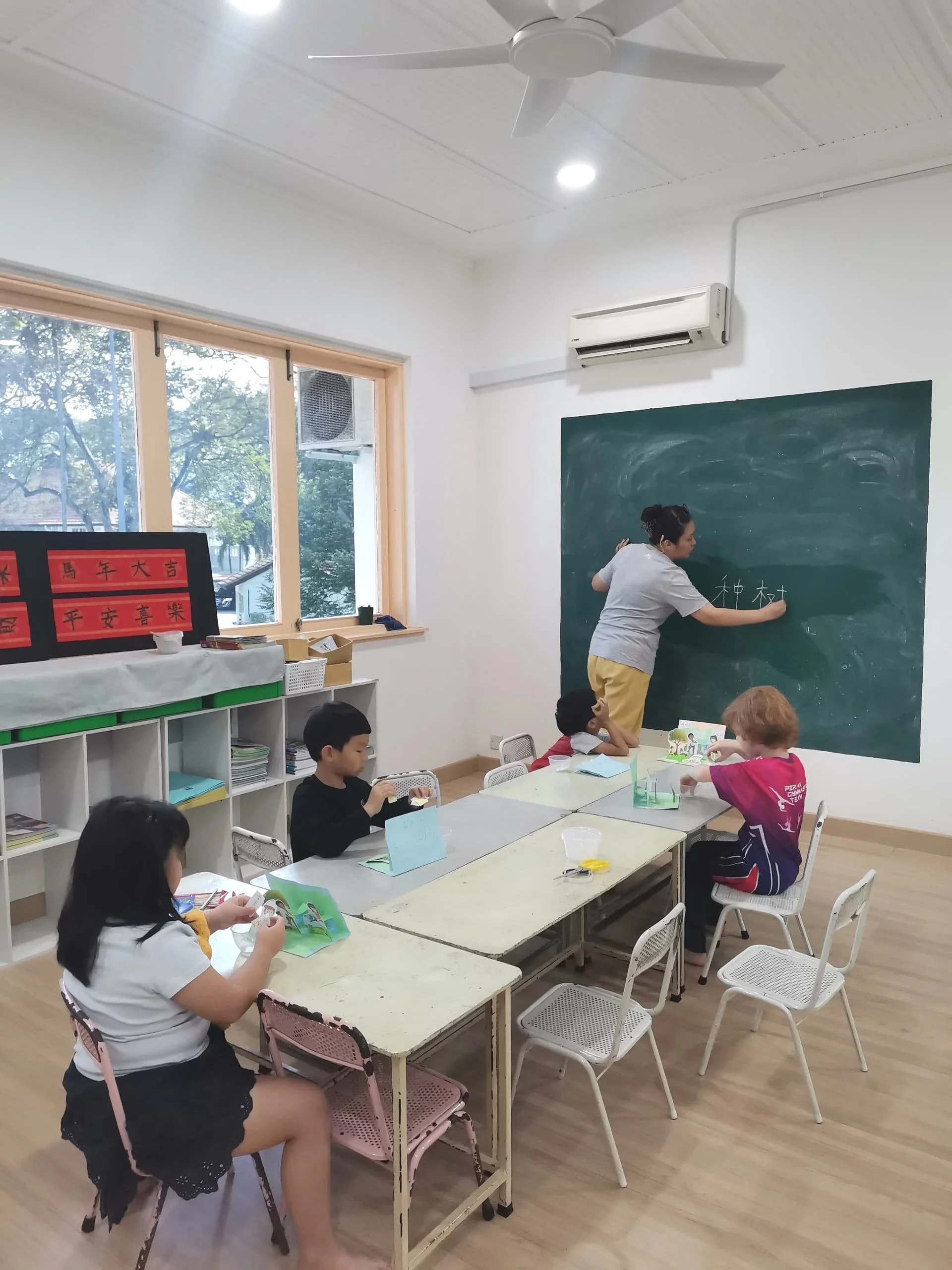 A young female teacher writing on a blackboard in a classroom with children seated at tables, some reading books. The classroom has large windows with trees outside, a ceiling fan, and an air conditioning unit.