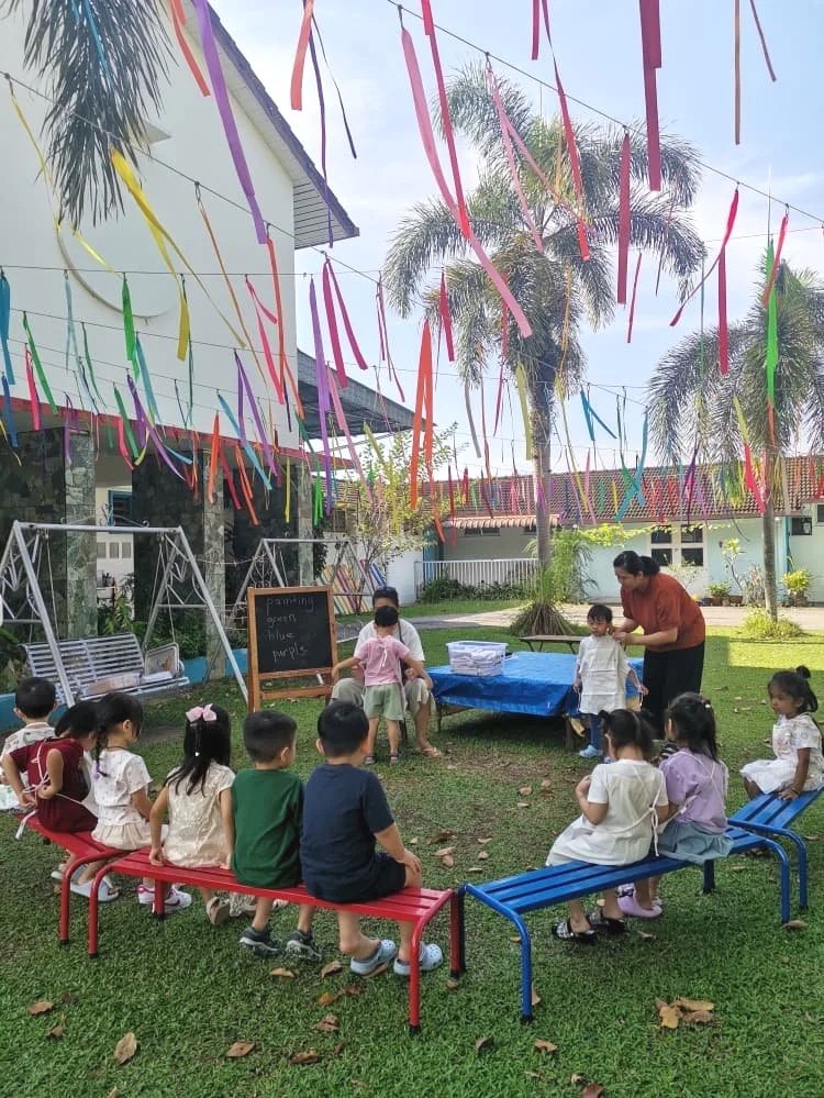 Children participating in an outdoor classroom or activity with an adult teacher, sitting on colorful benches, with decorations of colorful ribbons hanging overhead, and palm trees in the background.