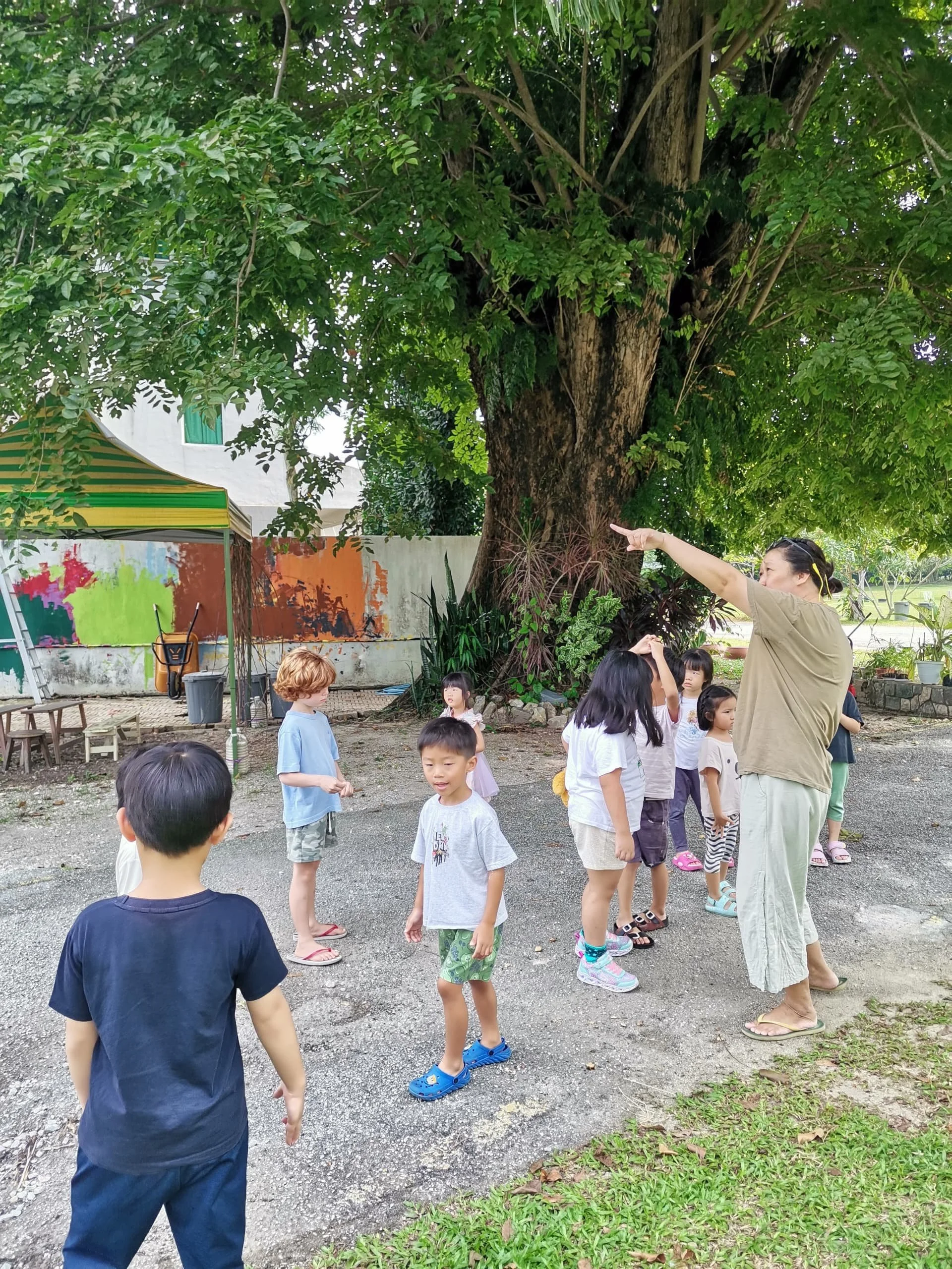 A woman appears to be instructing a group of children outdoors near a large tree, with a colorful tent and a mural in the background, on a gravel surface surrounded by greenery.
