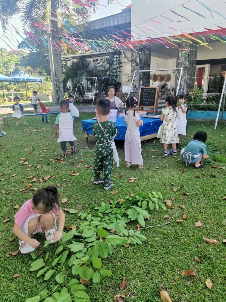 Children and a woman gather for an outdoor class or activity under a tent decorated with colorful streamers. Some children are wearing aprons, some are standing and some are sitting on the grass, with one girl crouching by a leafless branch in the fo