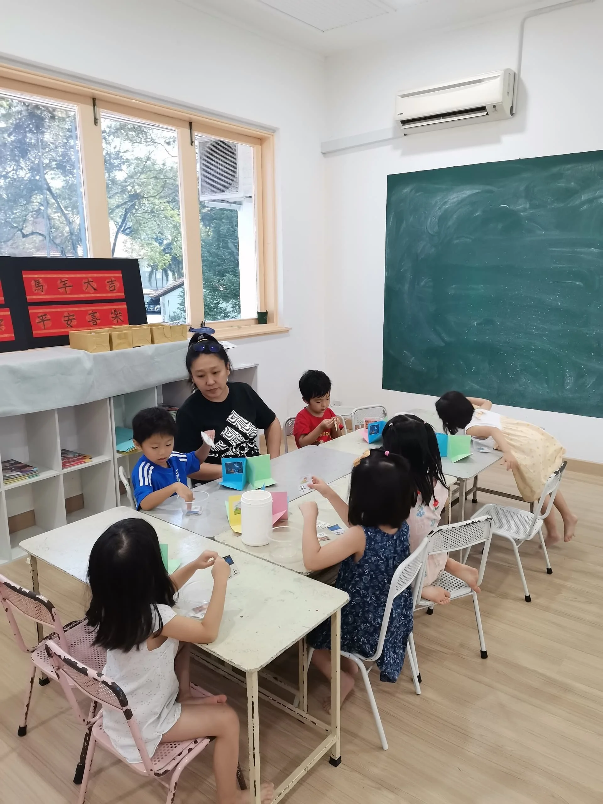Children and an adult sitting at tables in a classroom, engaged in activities, with a chalkboard on the wall and a window showing outside trees.