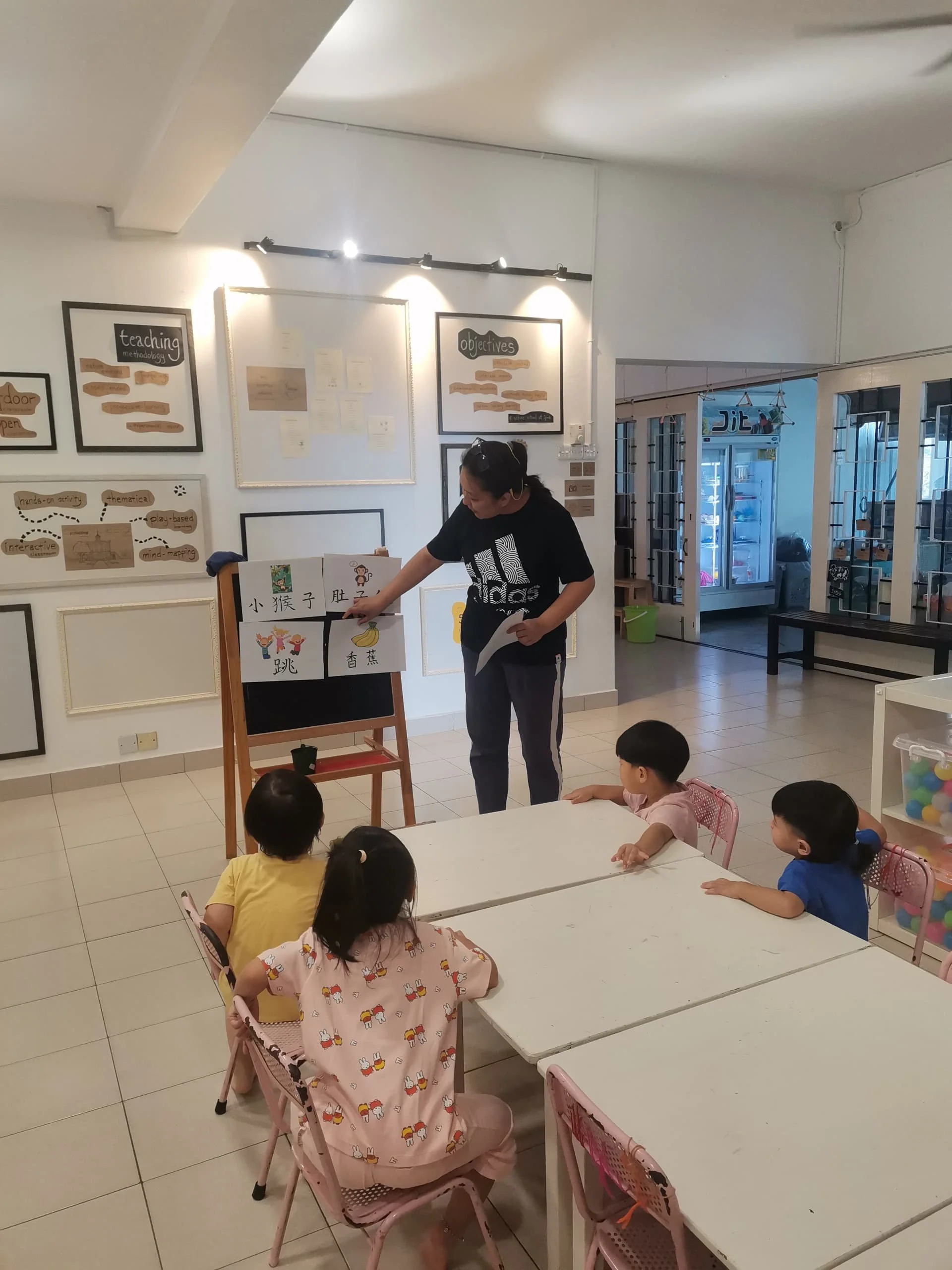 A woman teaching Chinese characters to young children in a classroom, with children seated around a white table and educational posters on the wall.