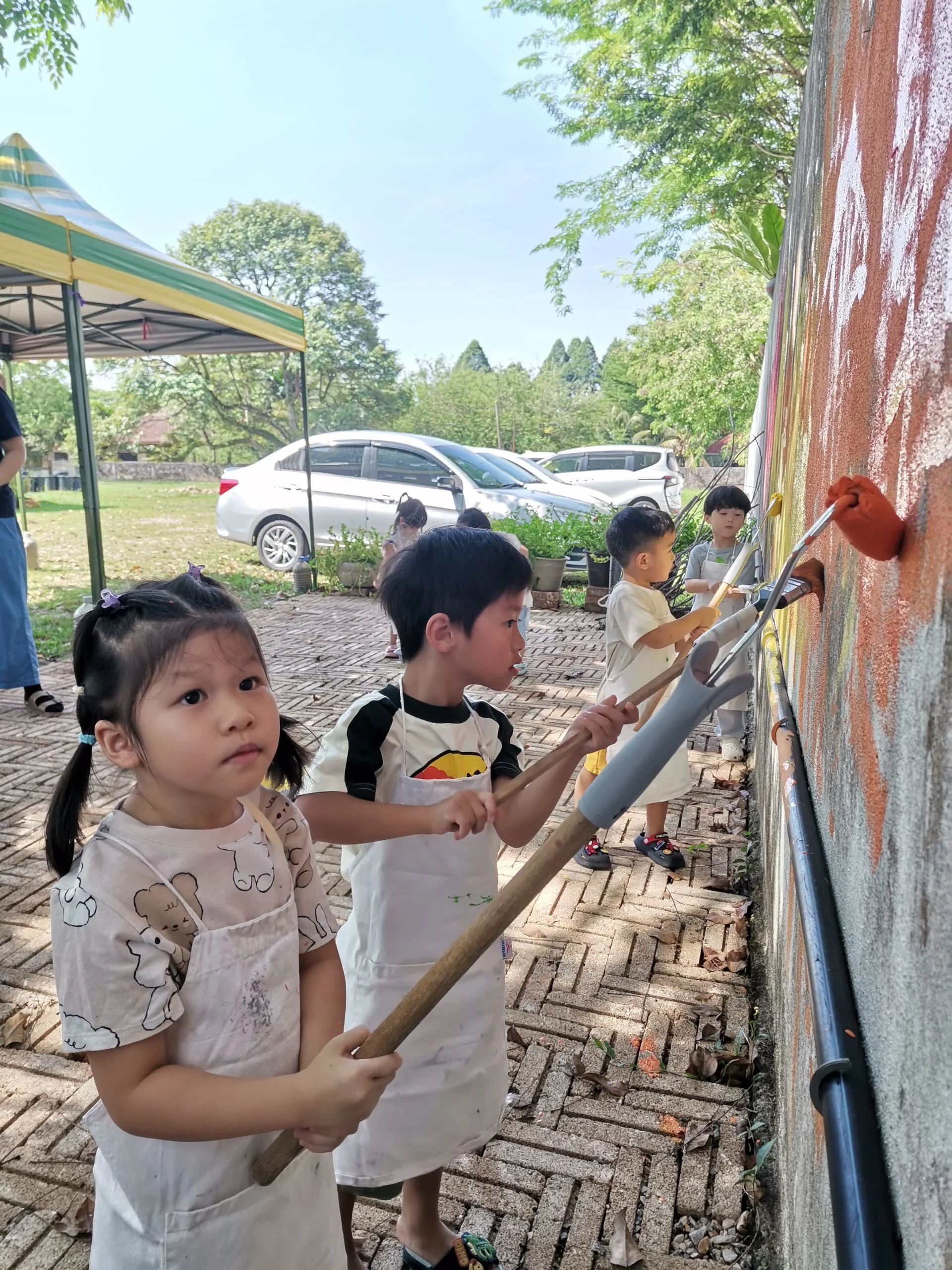 Children painting on an outdoor wall using brushes and roller sticks, with cars and trees in the background.
