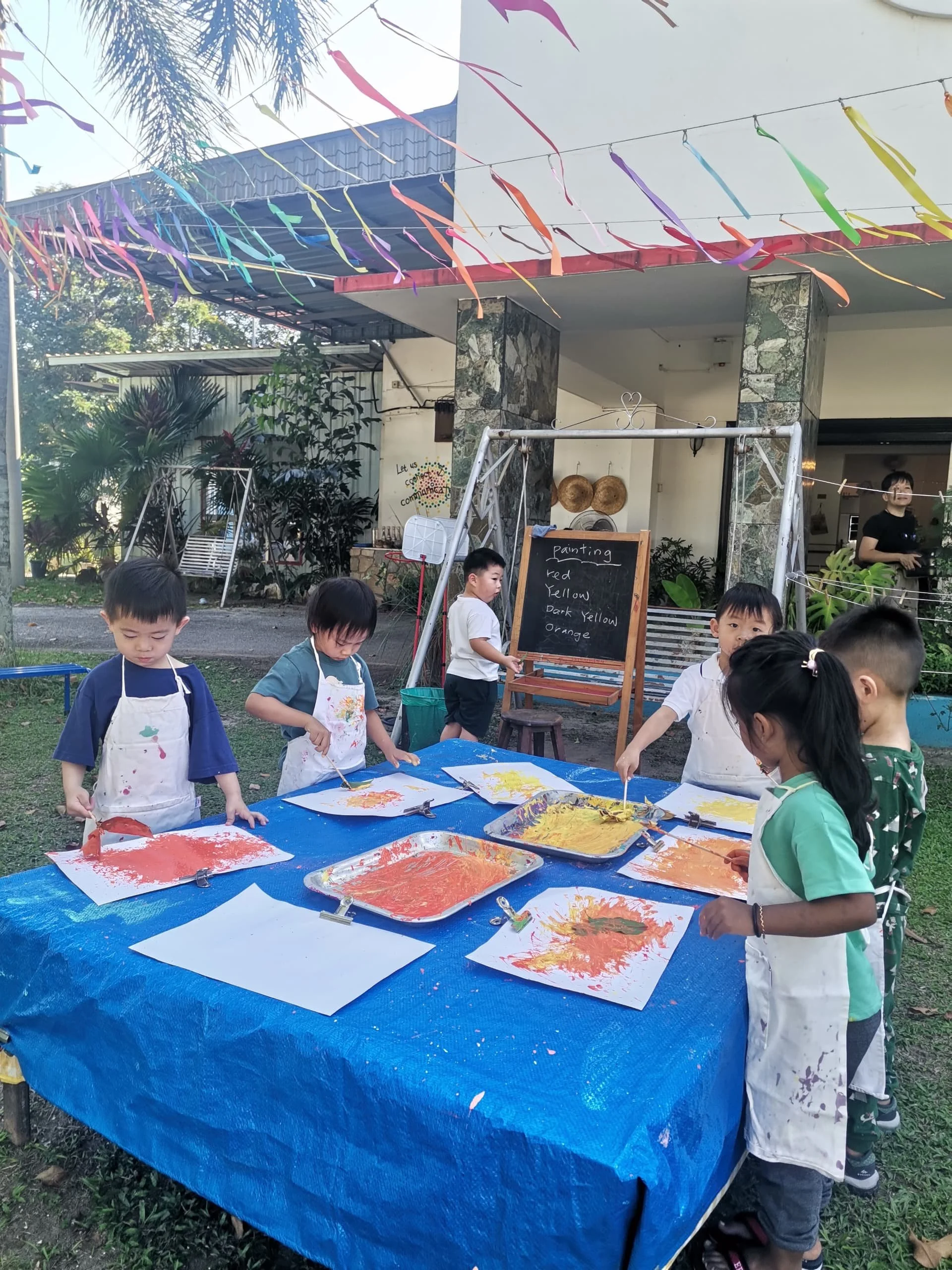 Children participating in outdoor painting activity, creating colorful artwork on paper with brushes, under a canopy decorated with multicolored streamers, in a garden setting with trees and plants.