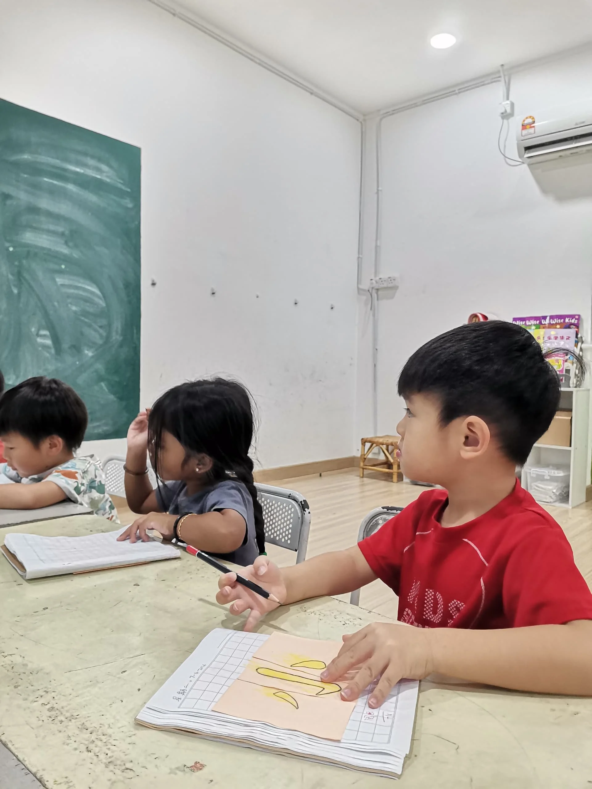 Children sitting at a classroom table, engaged in learning activities, with notebooks and writing materials in front of them.