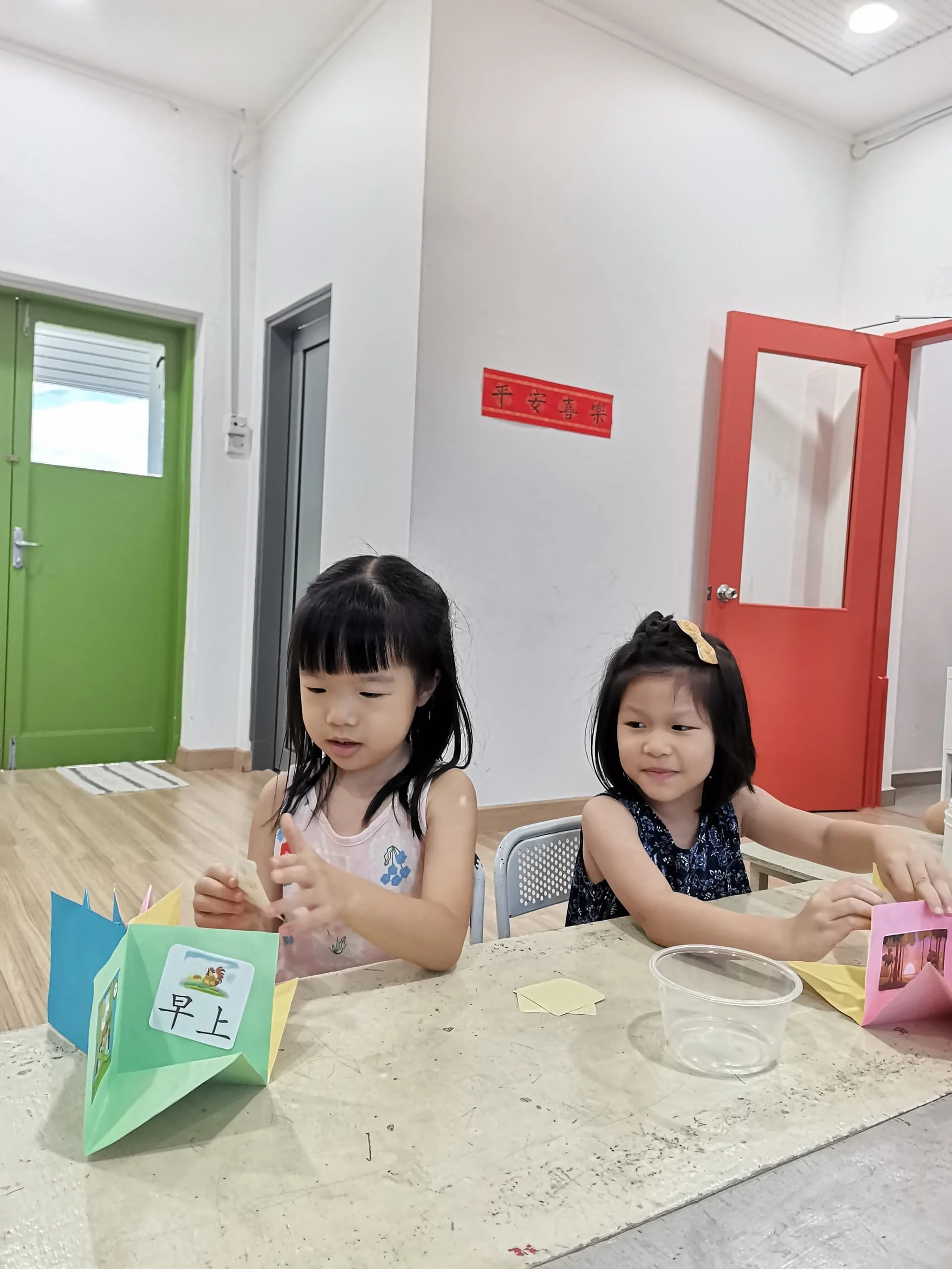 Two young girls sitting at a table with colorful origami costumes in front of them, inside a room with white walls, green and red doors, and a red banner with Chinese characters.