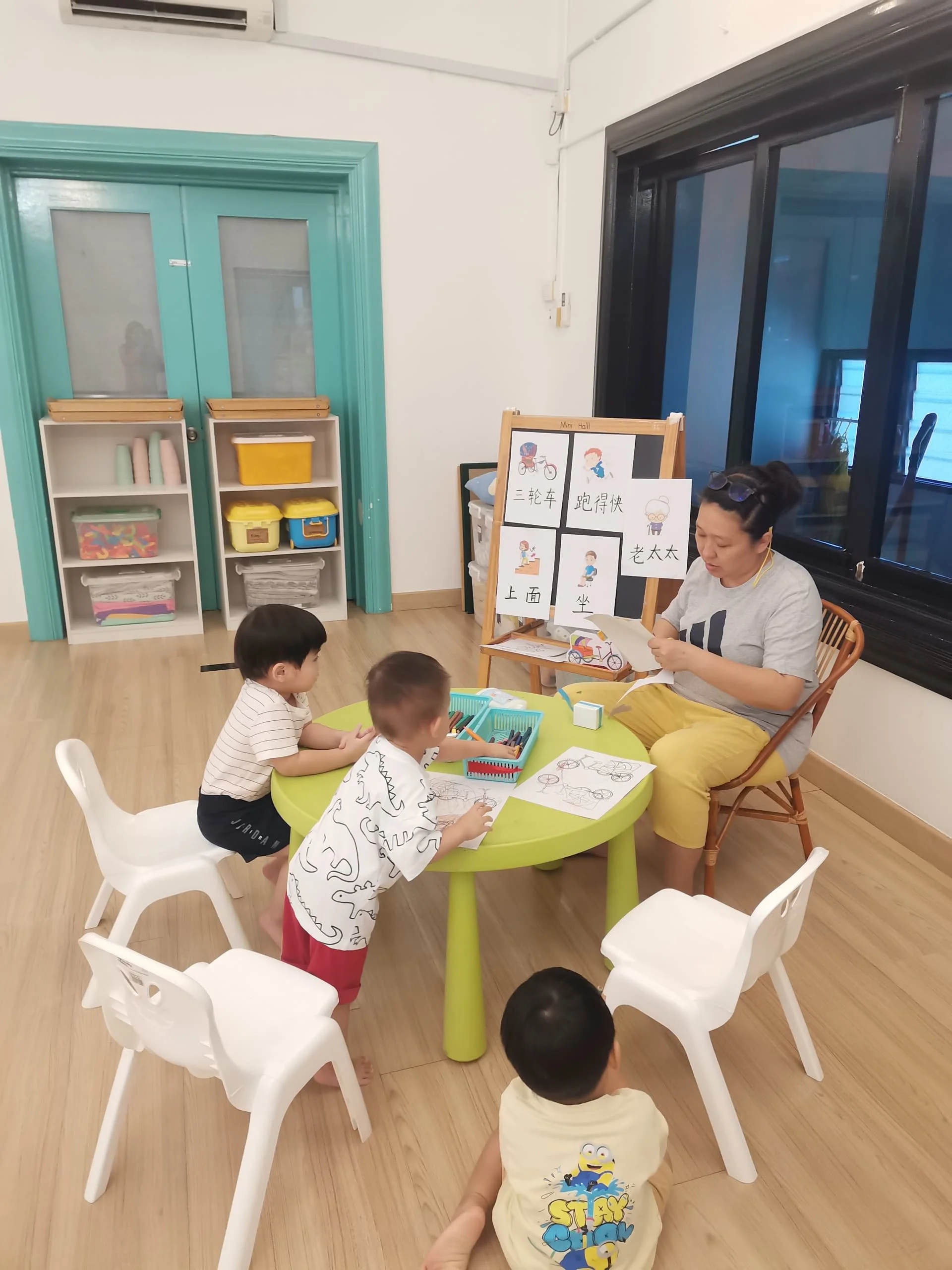 Children sitting around a green table holding coloring pages, with a woman showing a bike drawing to the children in a classroom setting that has shelves, charts with Chinese characters, and large windows.