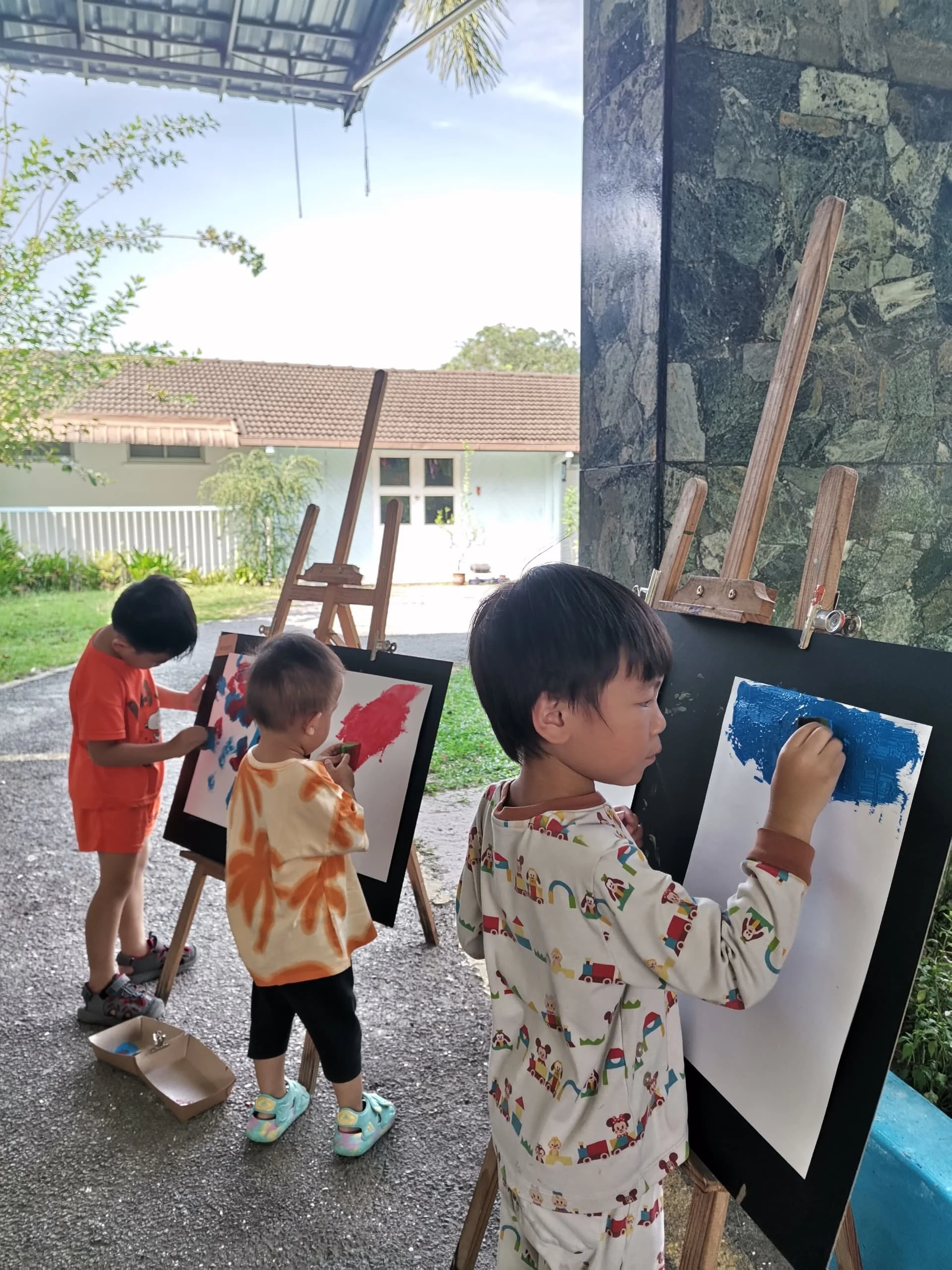 Children painting on canvases outdoors, standing and observing their artwork.