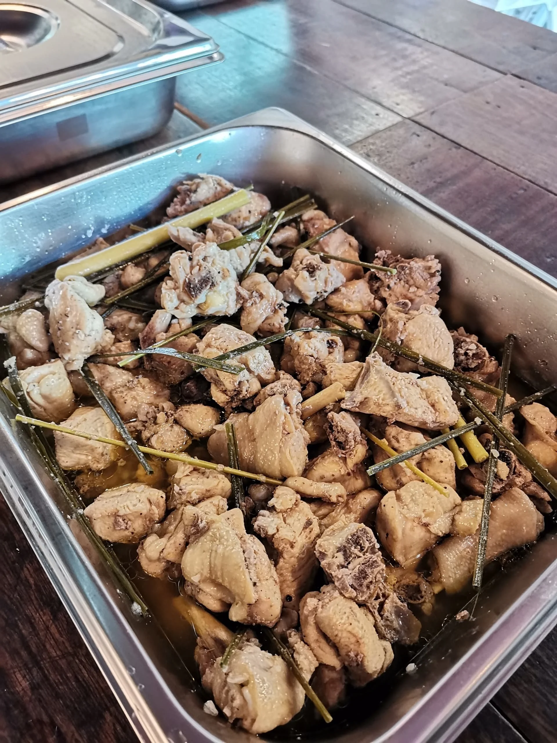 Chunks of cooked chicken with some herbs and seasonings in a metal container, with a wooden surface underneath.