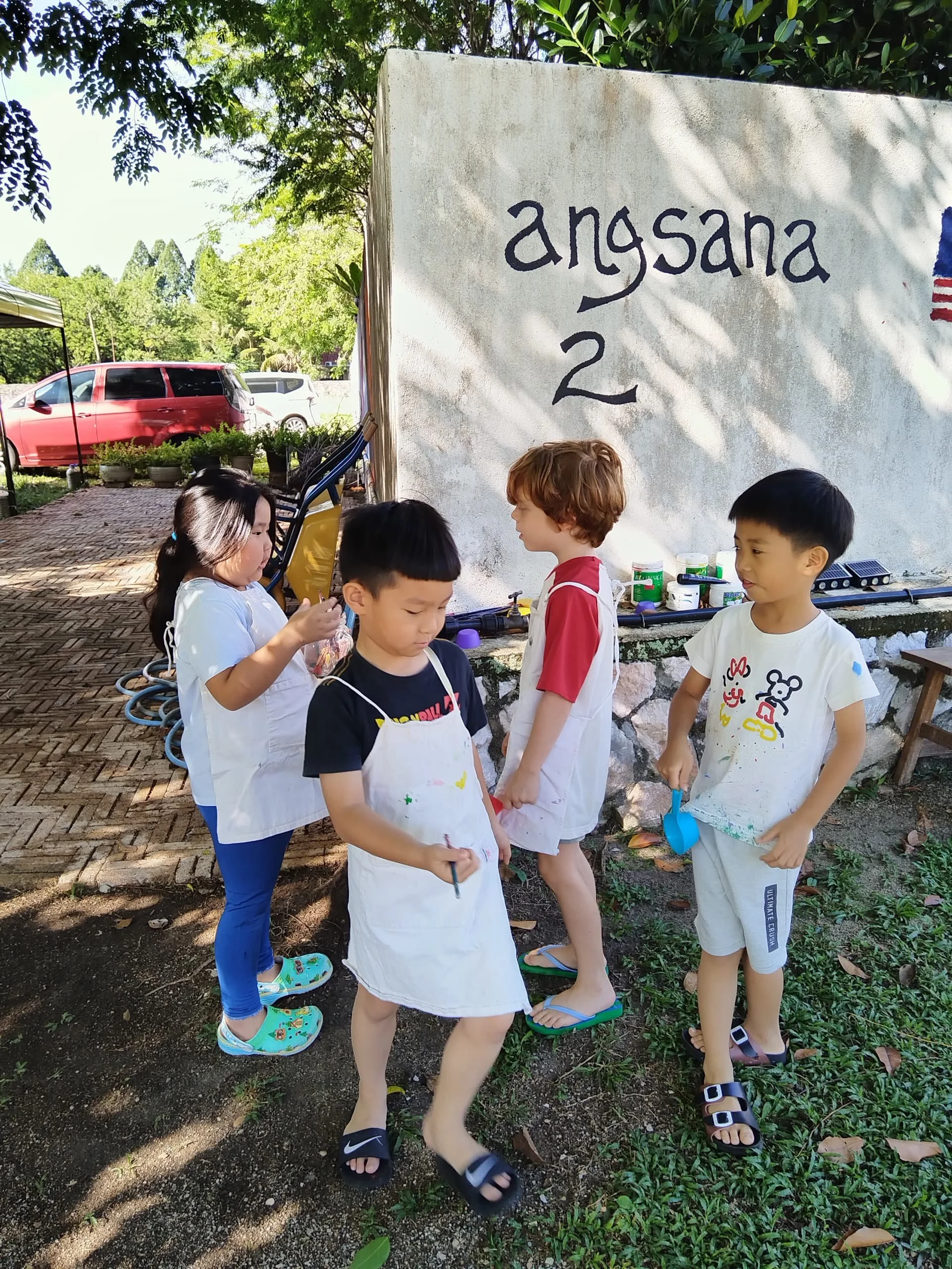 Group of children outdoors standing on a balance board, wearing white aprons, surrounded by trees and parked cars, with a sign that reads 'angasana 2' behind them.