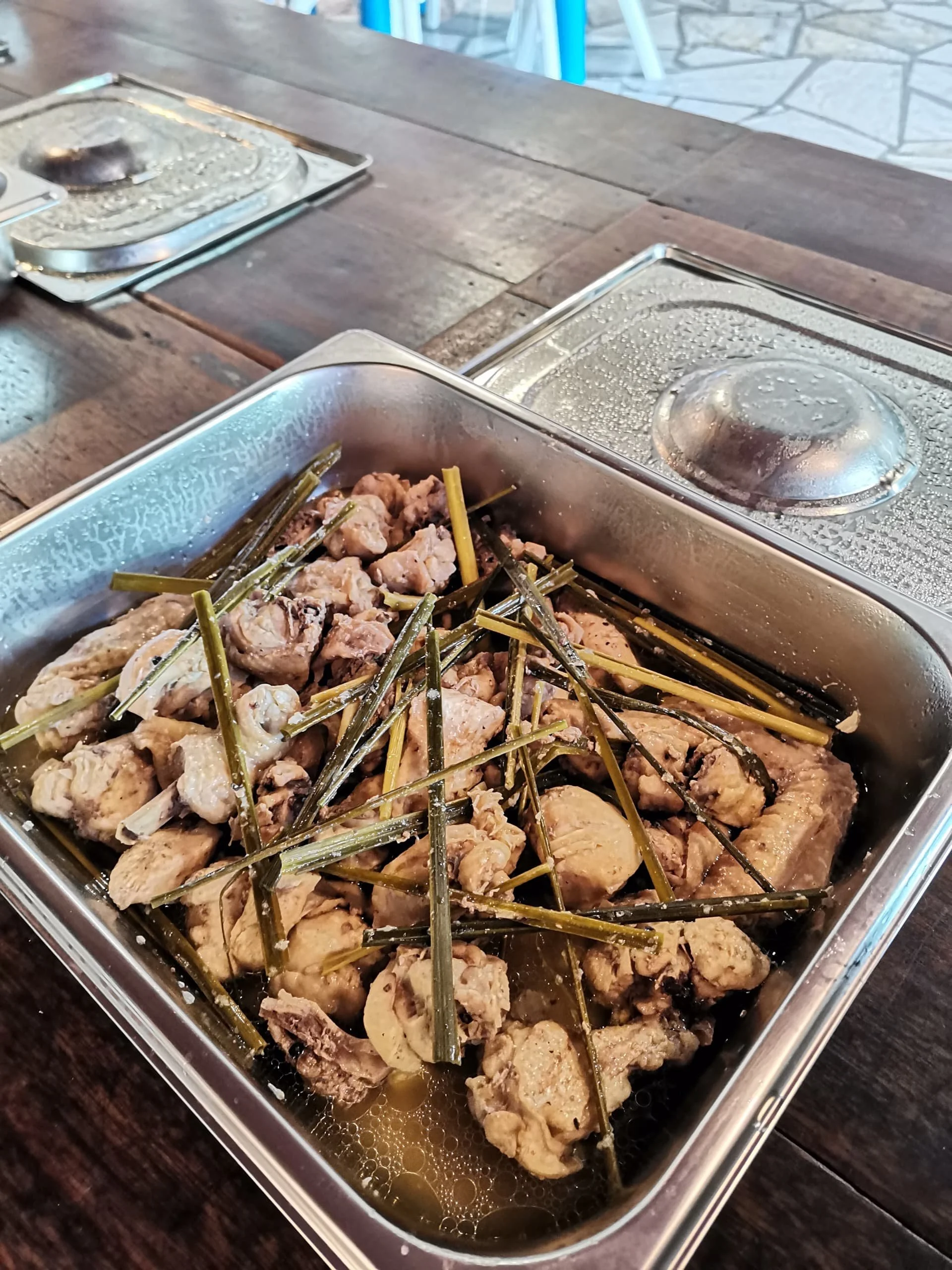 A close-up of a stainless steel serving tray with cooked chicken and green herbs in a broth, on a dark wooden table in a restaurant setting.