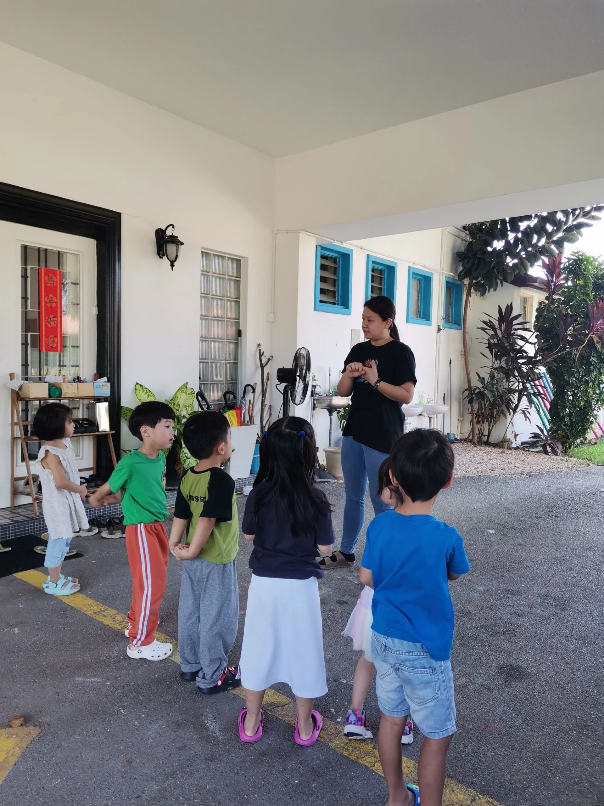 Children and a woman standing in a circle outside in a schoolyard or park, engaged in an activity. Young kids are wearing colorful casual clothing, and the woman is directing the activity. There are trees, a building with blue window frames, and vari