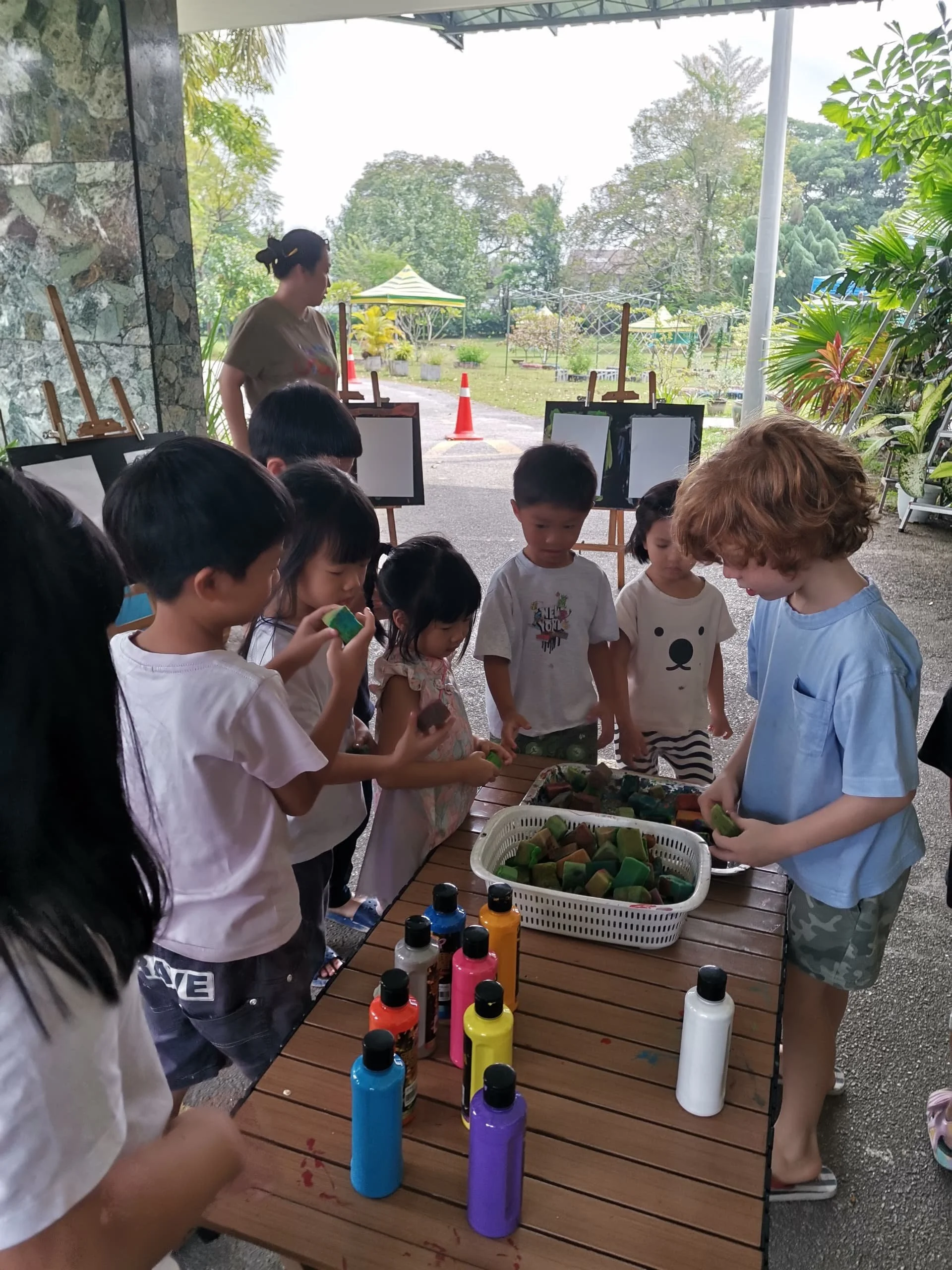 Children participating in a finger-painting activity outside, with colorful paint bottles on a wooden table, and a woman supervising in the background.