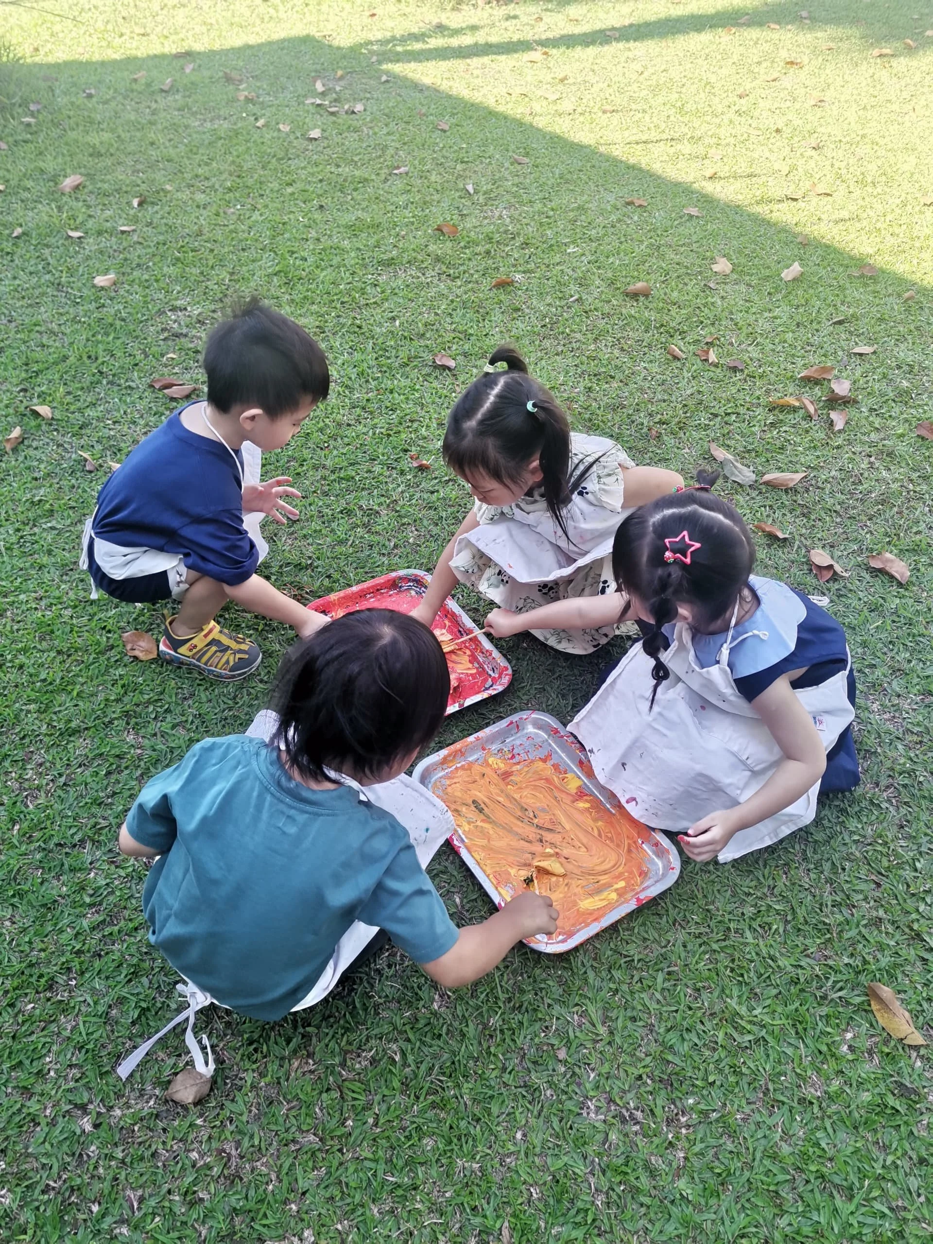 Children playing together on the grass, with trays of paint in front of them, engaging in an outdoor art activity.