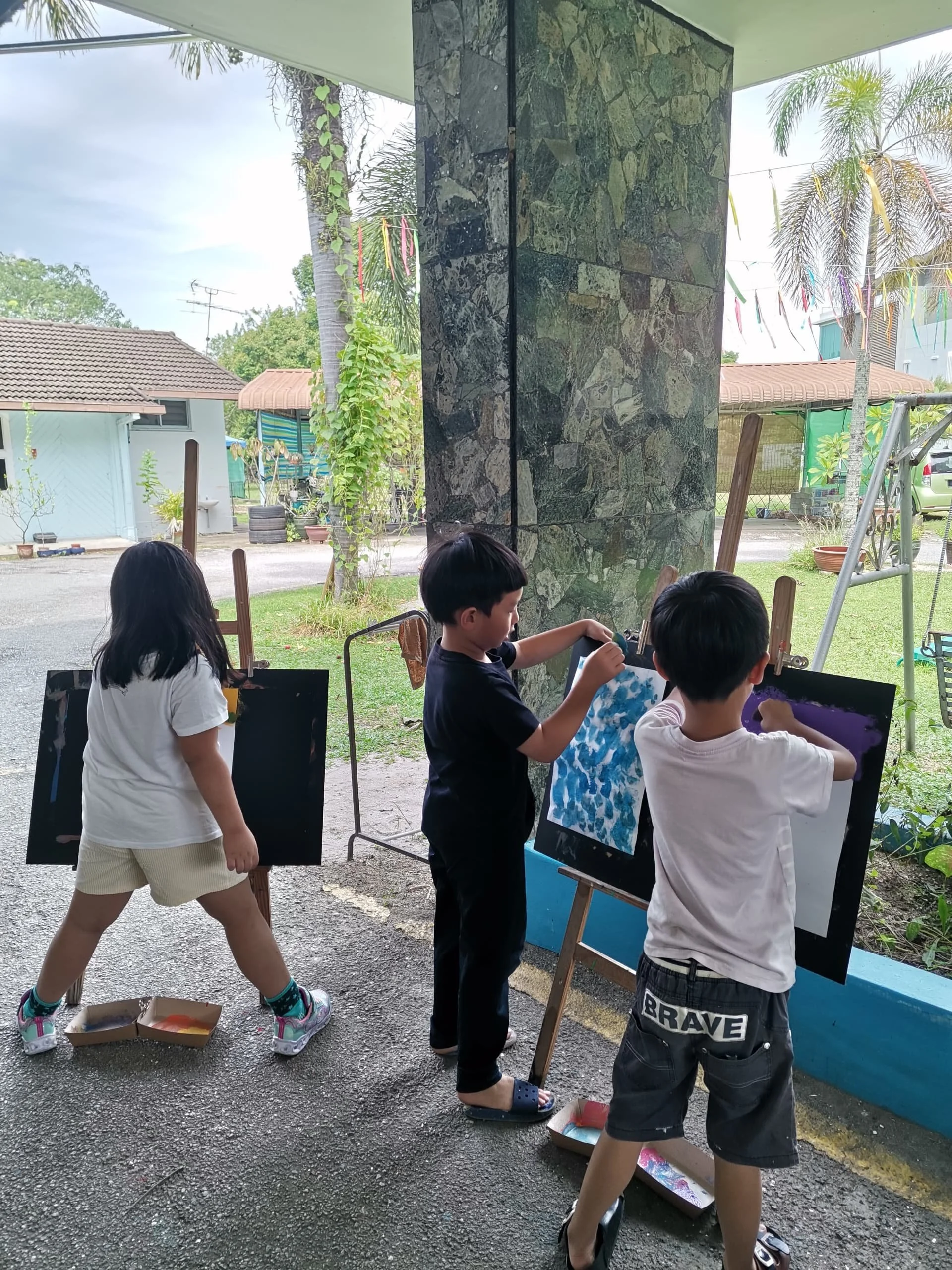 Children painting on canvases set on easels outdoors near a large stone pillar with trees and houses in the background.