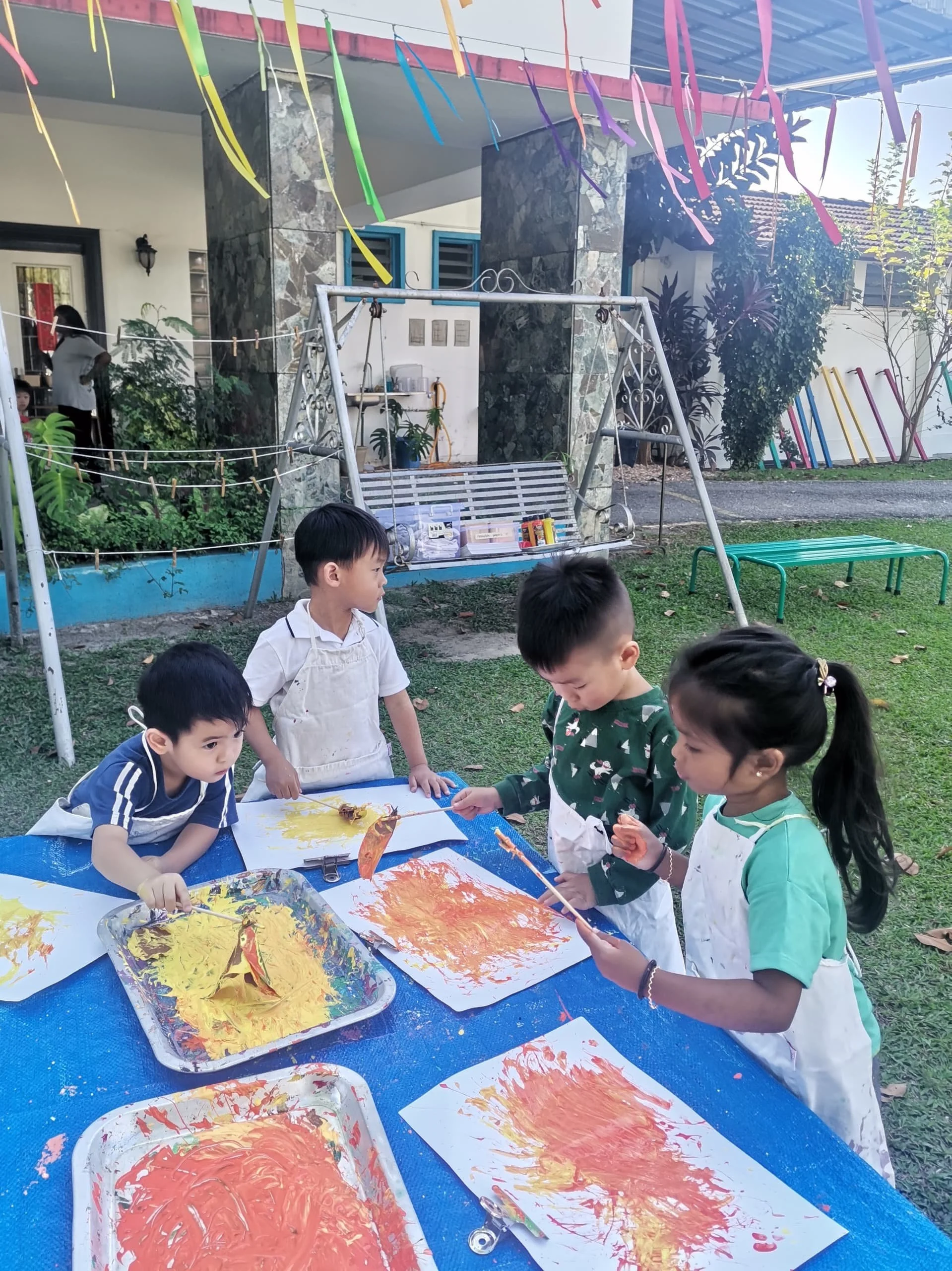 Children engaging in outdoor painting activity, with art supplies and colorful paintings on paper, set in a backyard with swing set, plants, and house in the background.