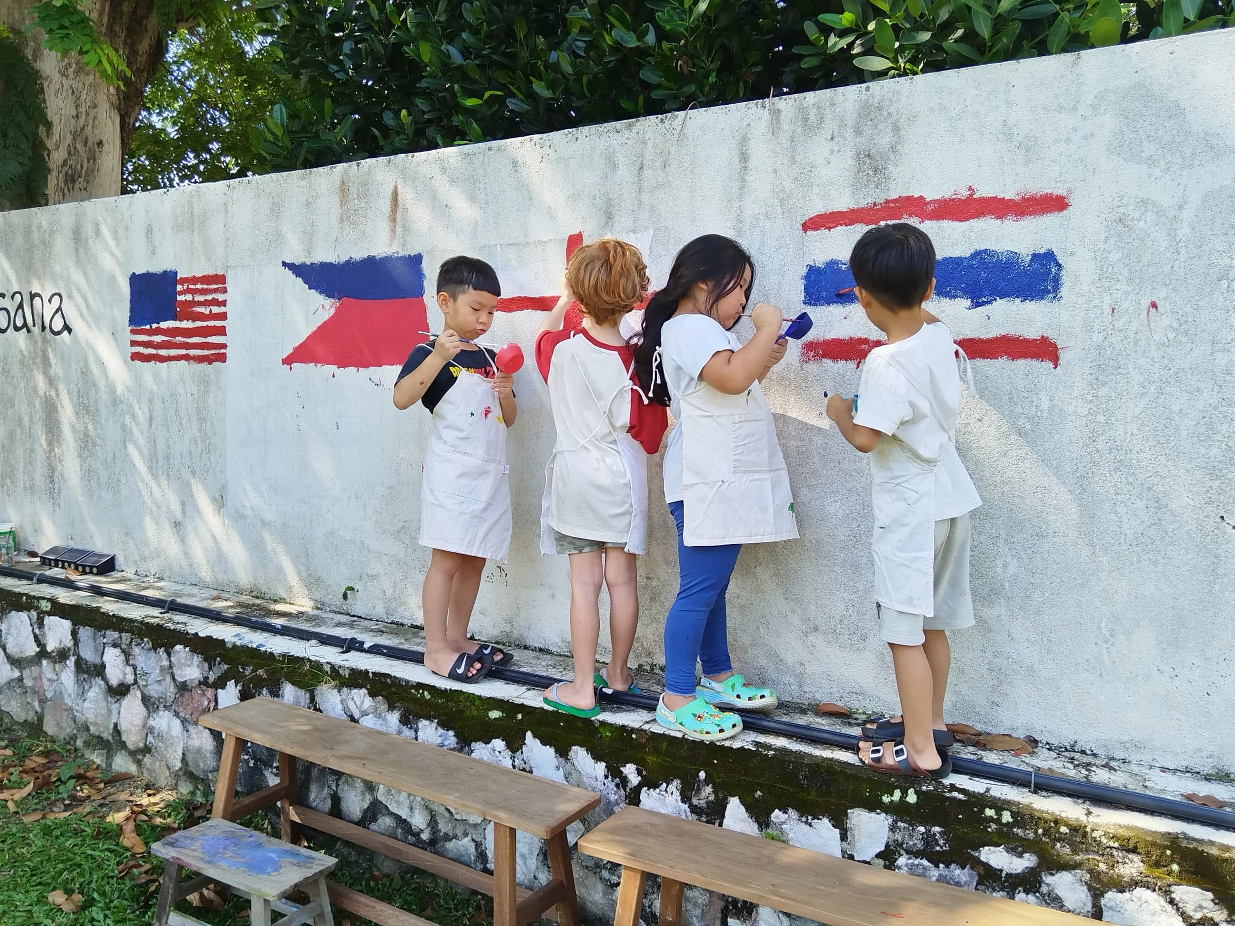 Five children standing on a narrow concrete ledge, painting flags of the United States and France on a white wall outdoors, with trees and greenery in the background.