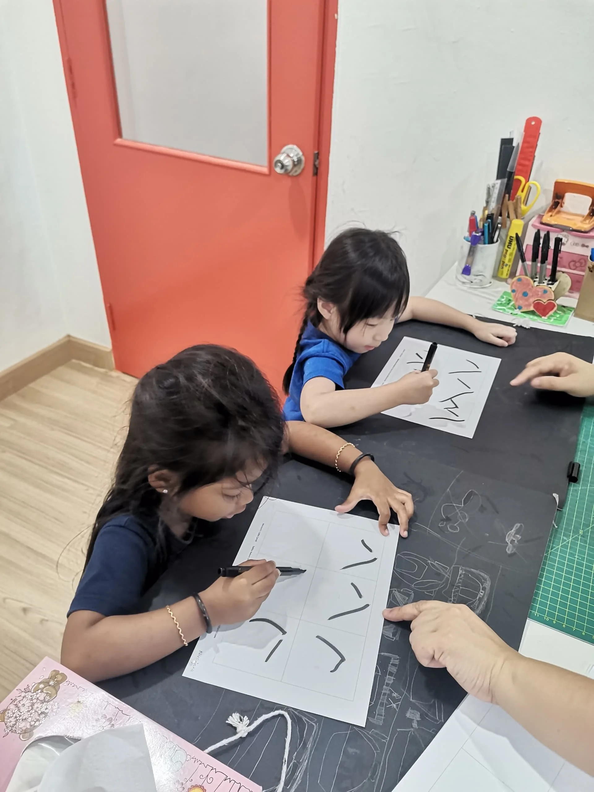 Children sitting at a table, drawing on sheets of paper with black markers. Various art supplies and craft materials are on the table, including a container with pens and scissors, and decorative items. A woman is helping one of the children. The set