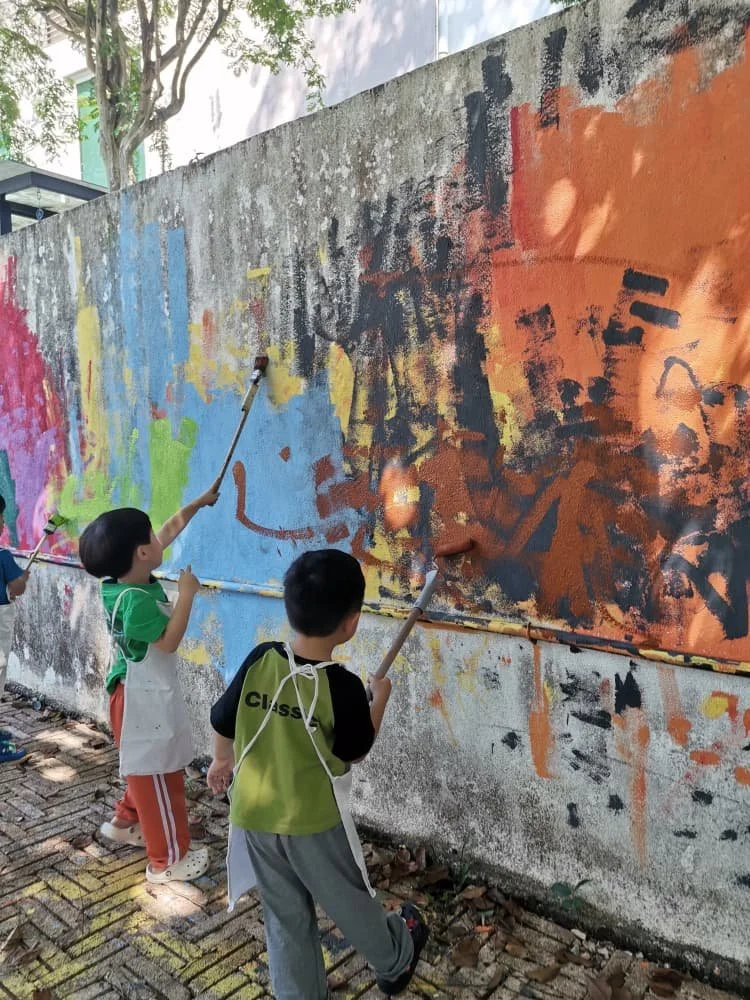 Three children are painting a colorful mural on a concrete wall outdoors, using paint rollers. The mural features a mix of bright colors and abstract patterns.