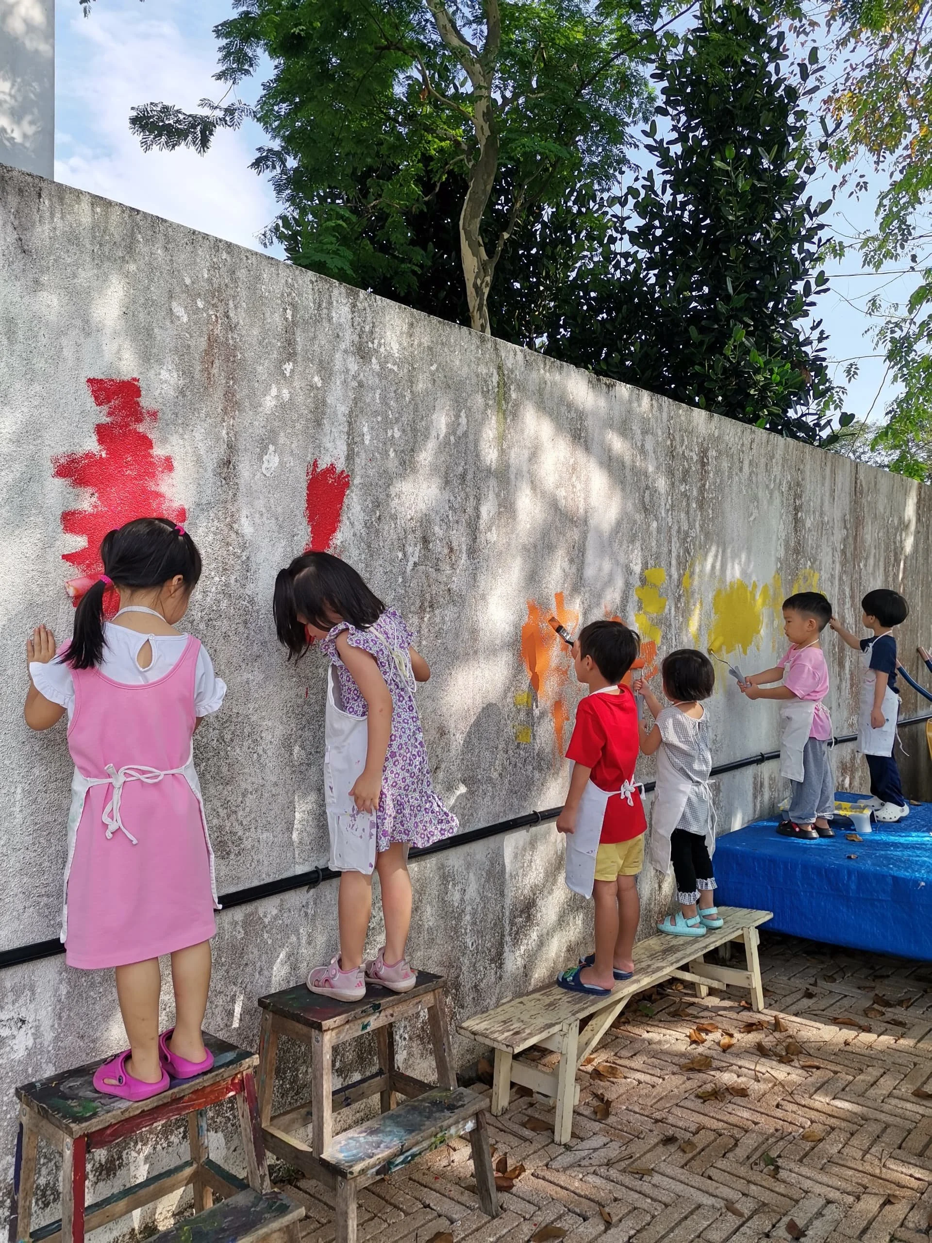 Children painting on a concrete wall outdoors, standing on small wooden platforms for support, wearing aprons and casual clothes, with trees and blue sky in the background.