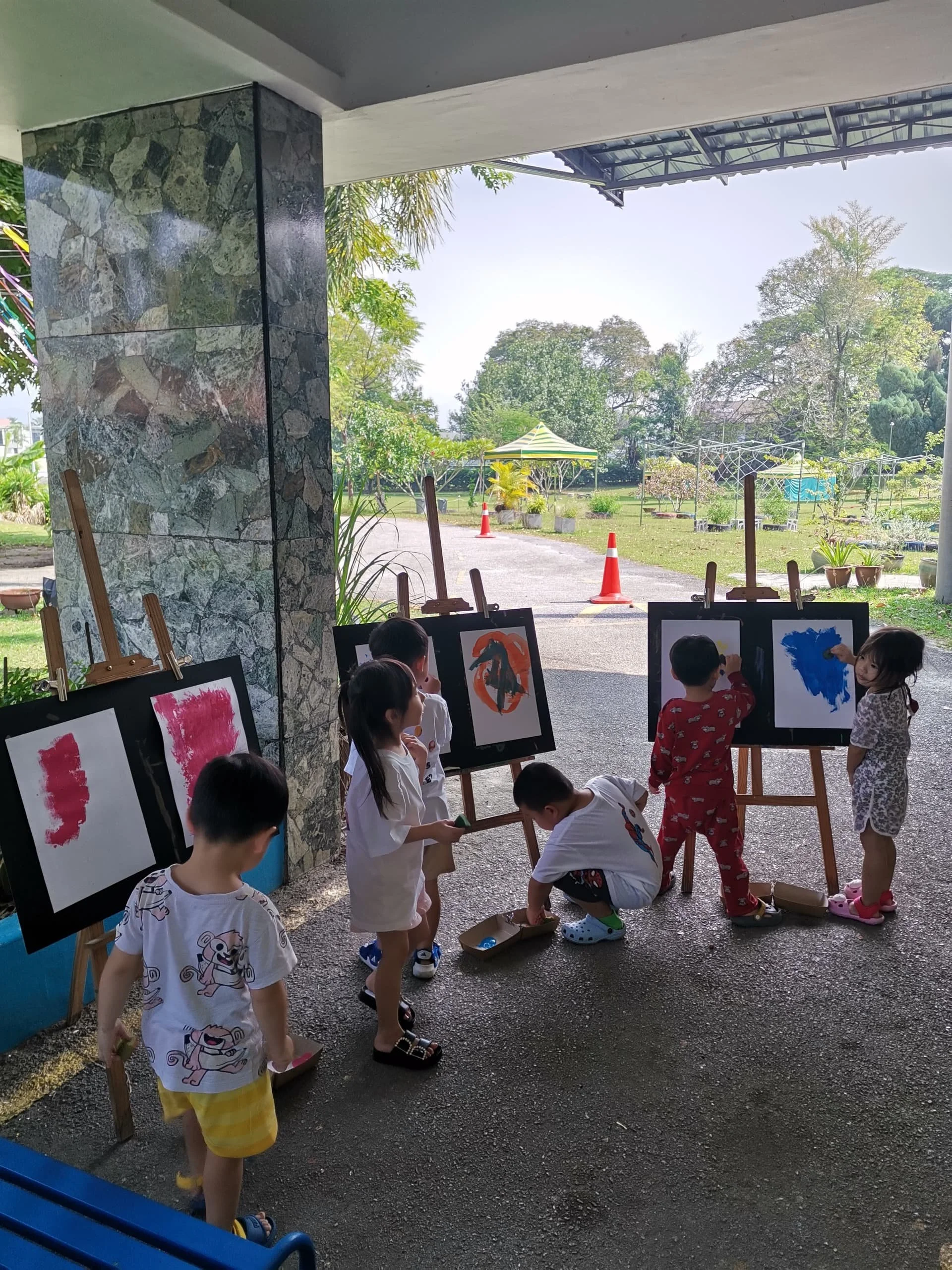 Children watching and drawing animal pictures outdoors in a shaded area with trees, potted plants, and orange traffic cones visible in the background.