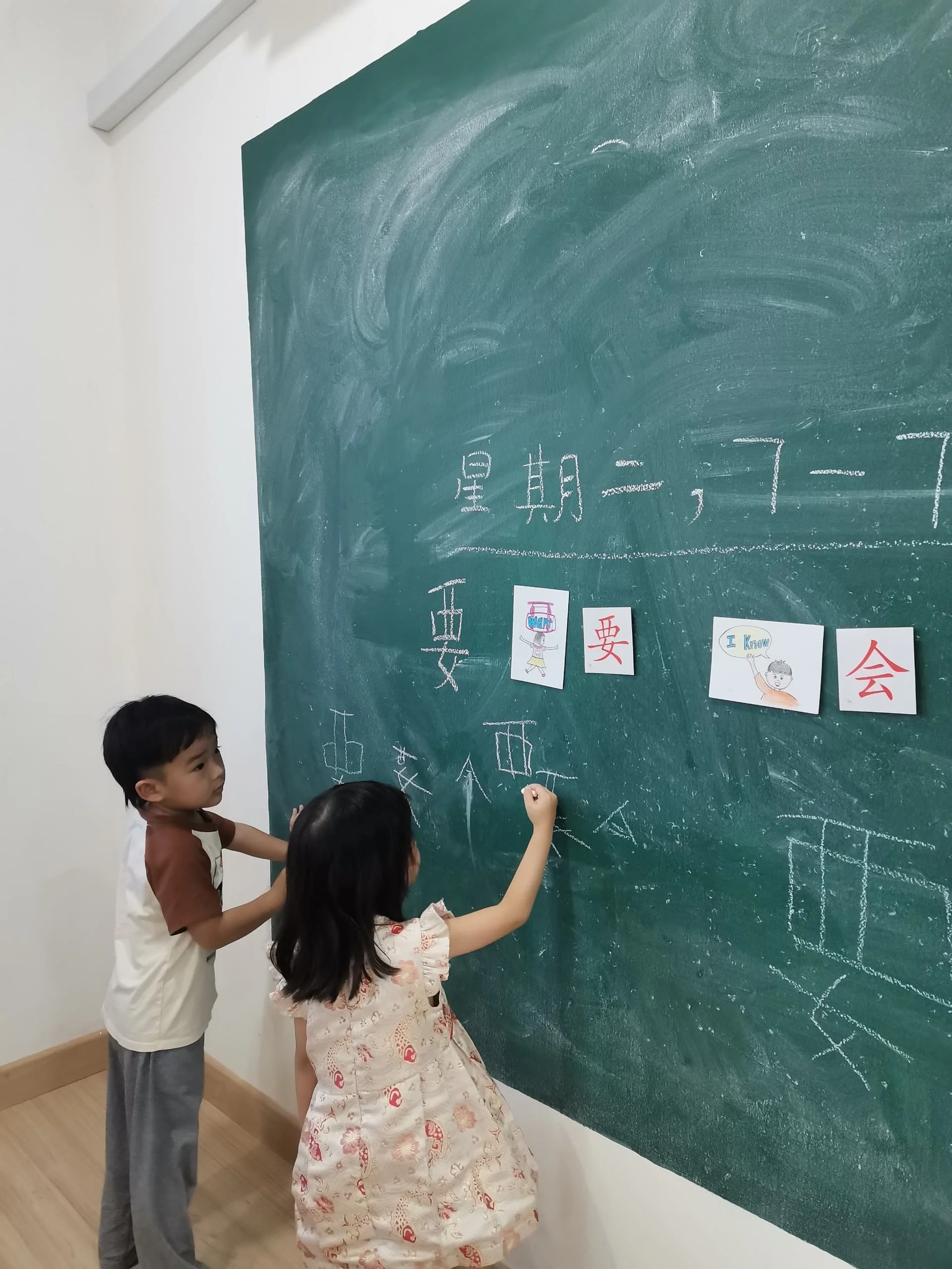 Two children, a boy and a girl, are standing at a large green chalkboard. The girl is drawing on the blackboard, and the boy is watching. There are Japanese characters and drawings on the chalkboard, including small pictures of children holding signs