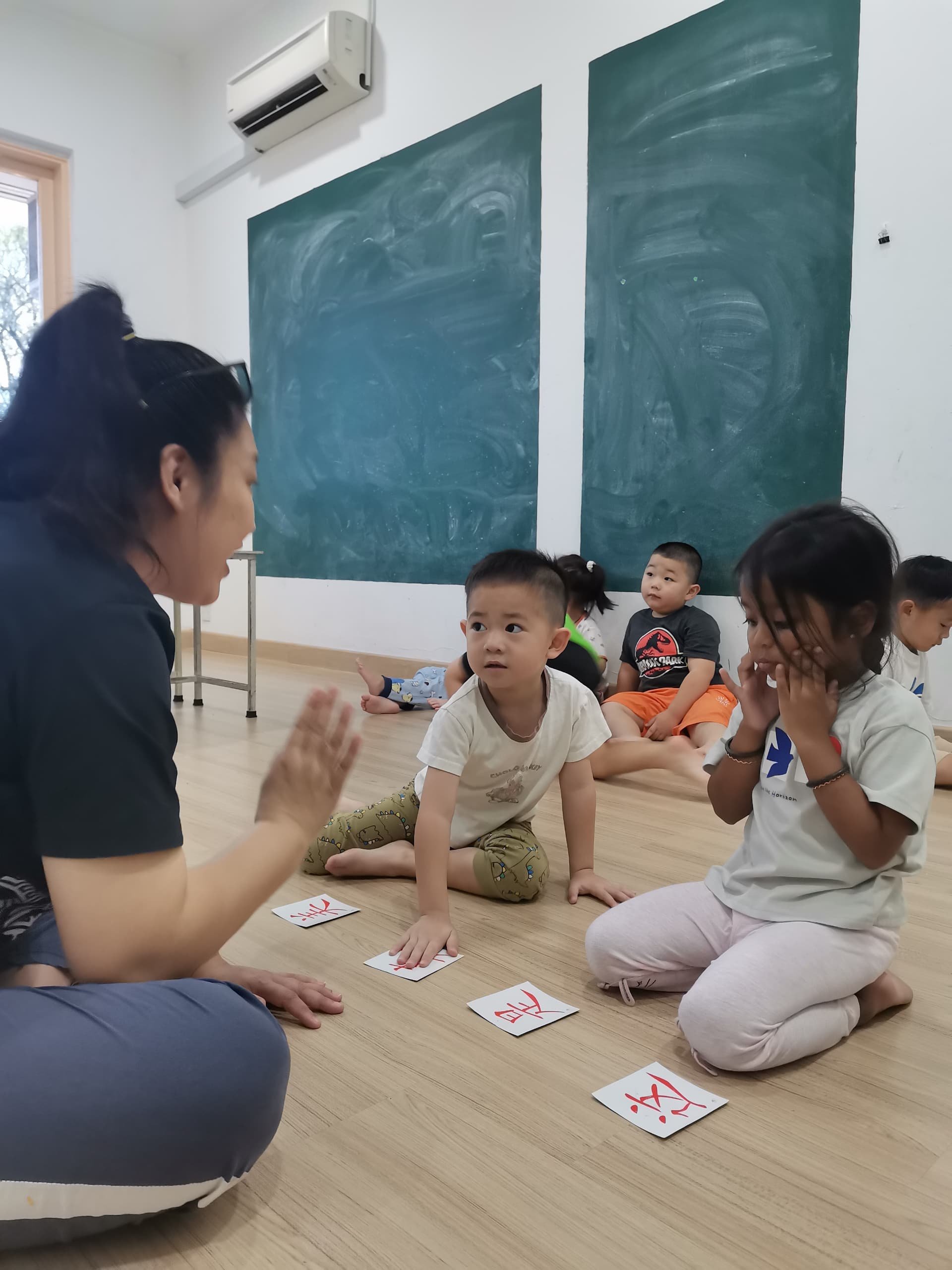 A group of children sitting on a wooden floor in a classroom with blackboards on the wall, engaging in an activity with a teacher, who is gesturing with her hands, and cards with words on the floor.