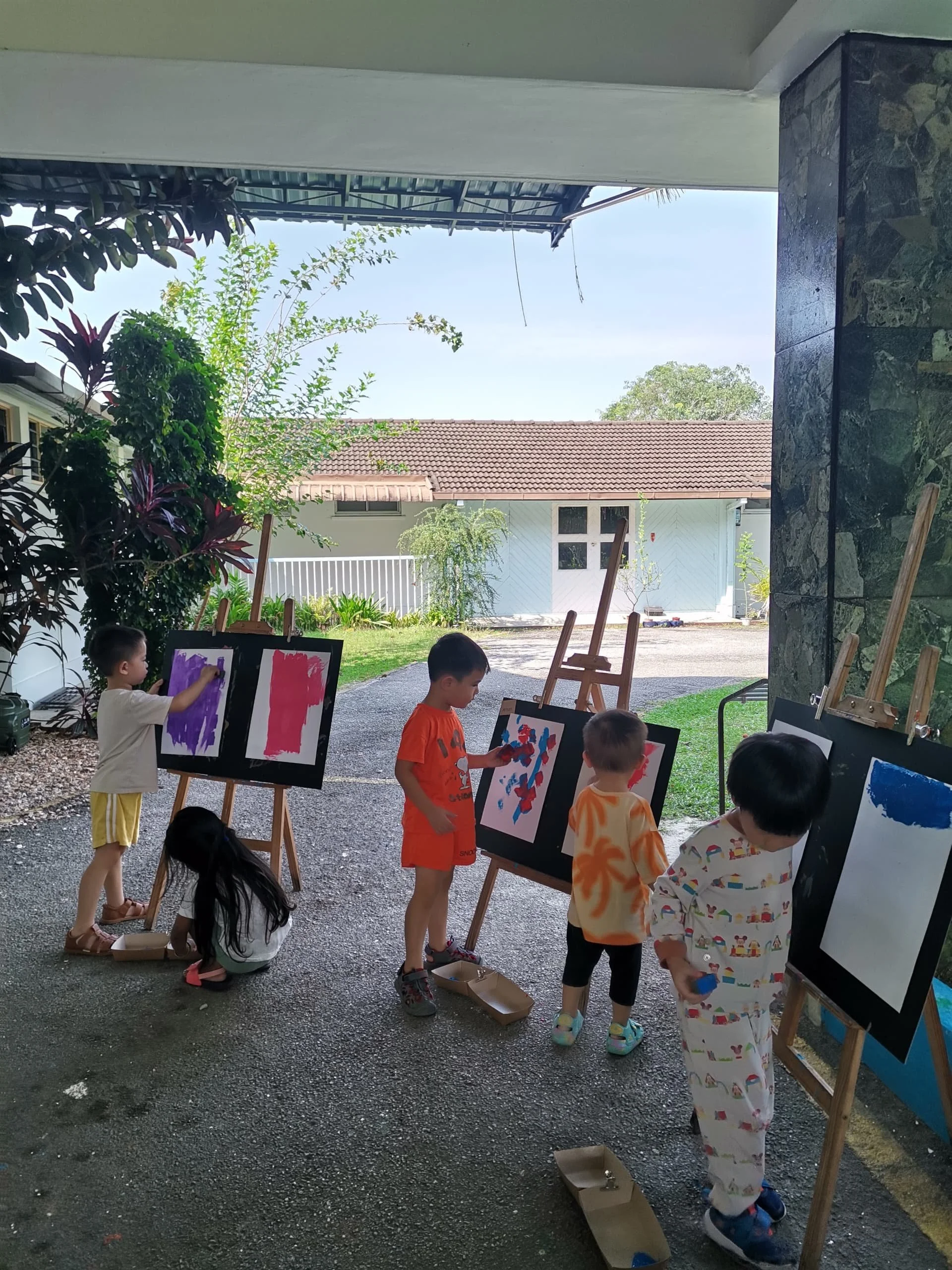 Children painting on canvases set on easels outdoors under a covered area, with a house and trees visible in the background.