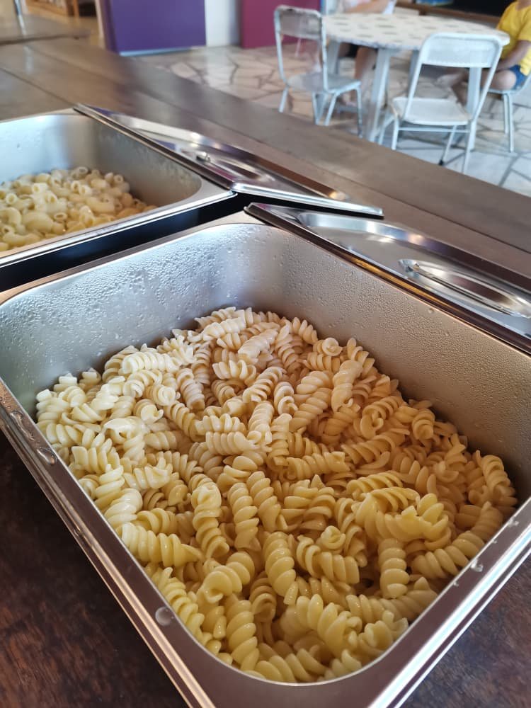 Two chafing dishes filled with cooked rotini pasta at a buffet table in a restaurant or cafeteria setting.