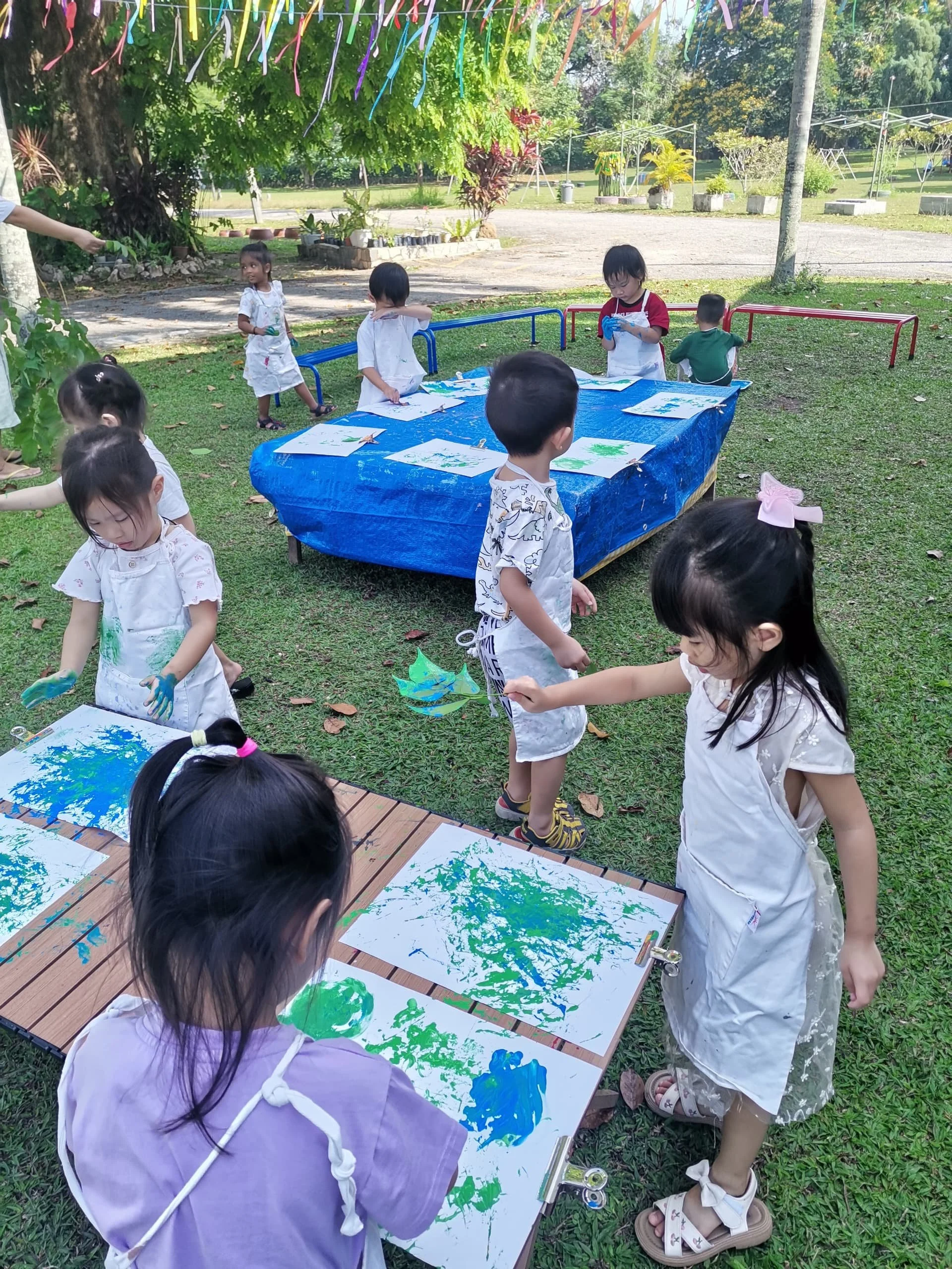 Children engaging in outdoor painting activity, some mixing colors on their papers, with trees and greenery in the background.
