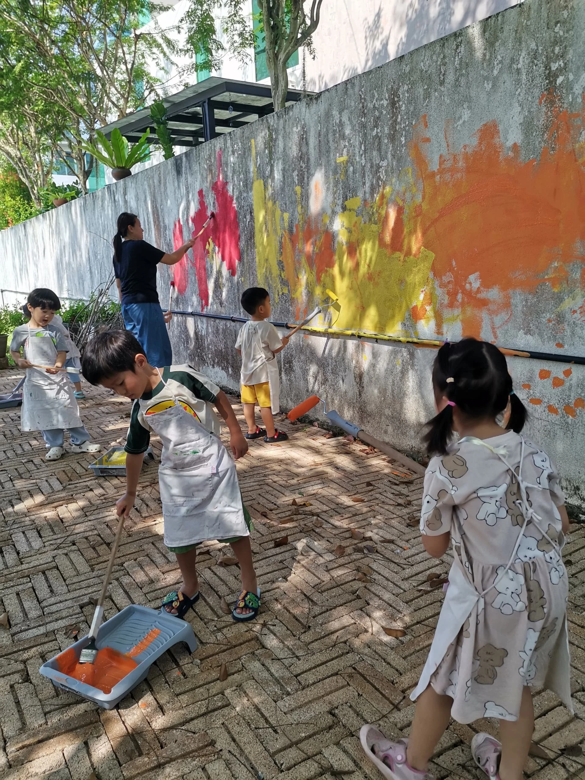 Children and an adult painting a large concrete wall outdoors. The children are wearing aprons and using paint rollers and brushes to create colorful artwork on the wall. There are trees and plants around, and the scene appears to be part of an outdo
