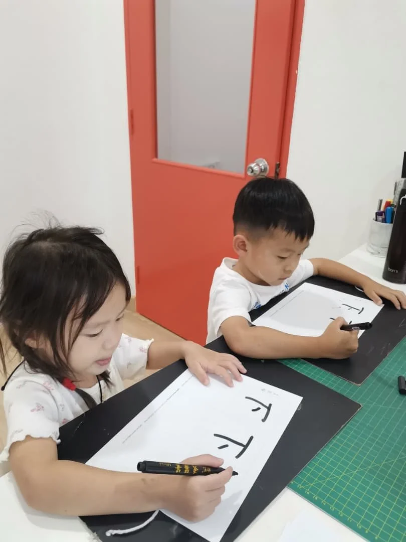 Two children sitting at a desk, practicing writing the letter 'T' on white sheets with black markers, in a classroom or study setting.