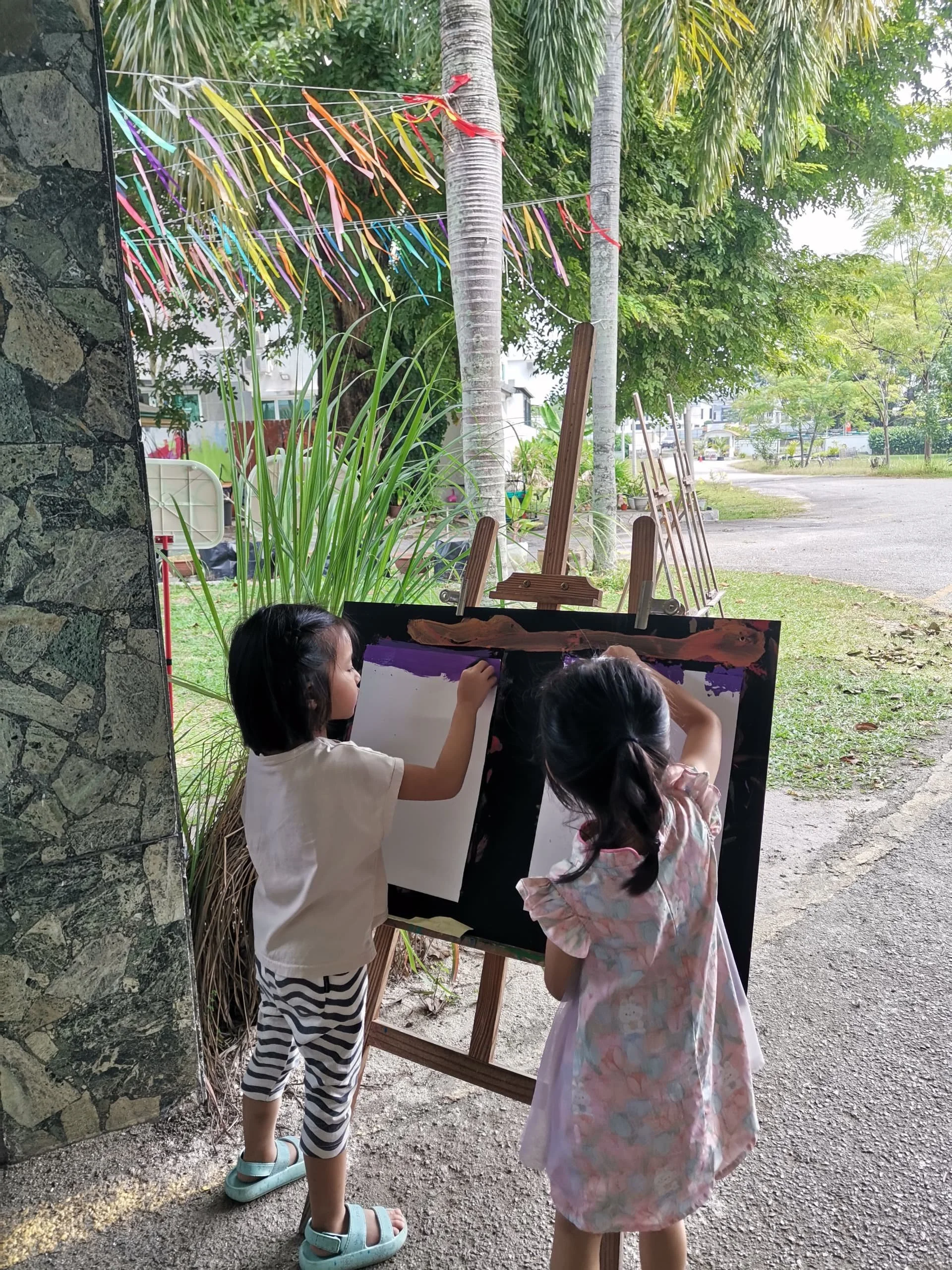 Two children, a girl and a boy, drawing on a large piece of paper outdoors next to a stone wall, with trees and colorful decorations in the background.