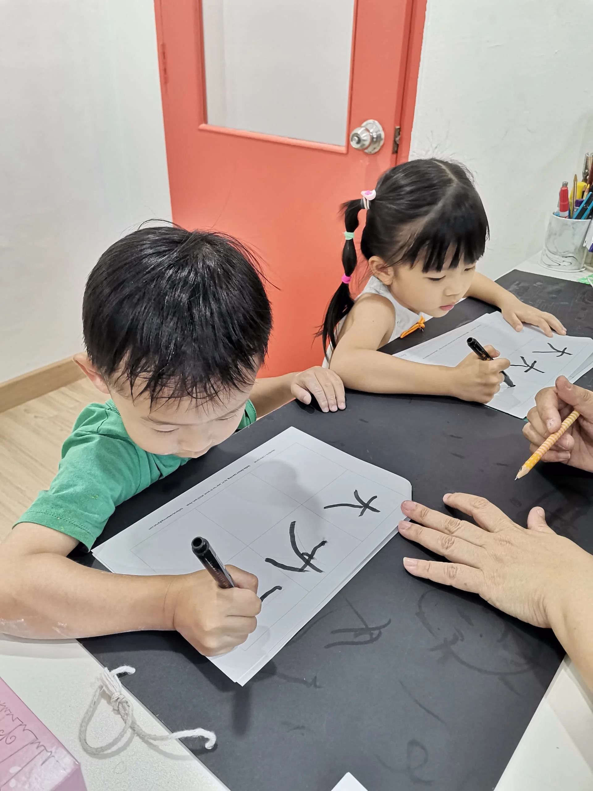 Two children, a boy and a girl, practicing writing Chinese characters on worksheets, with an adult guiding them, in a classroom setting.
