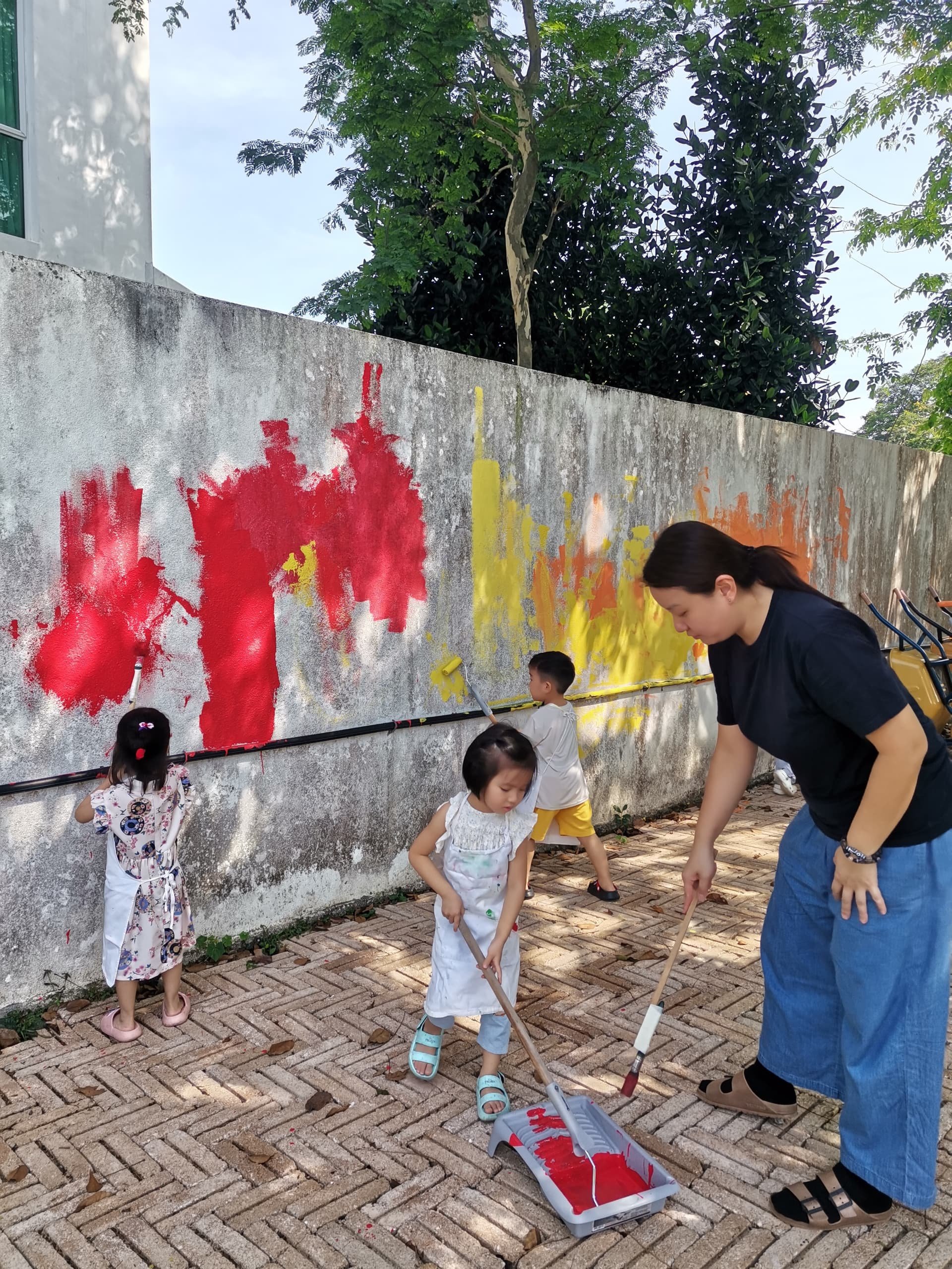 Children and an adult painting a mural on a concrete wall outdoors. The kids are using brushes and paint rollers, and some are wearing white clothes. The mural features red, yellow, and orange colors. Trees with green leaves are visible in the backgr
