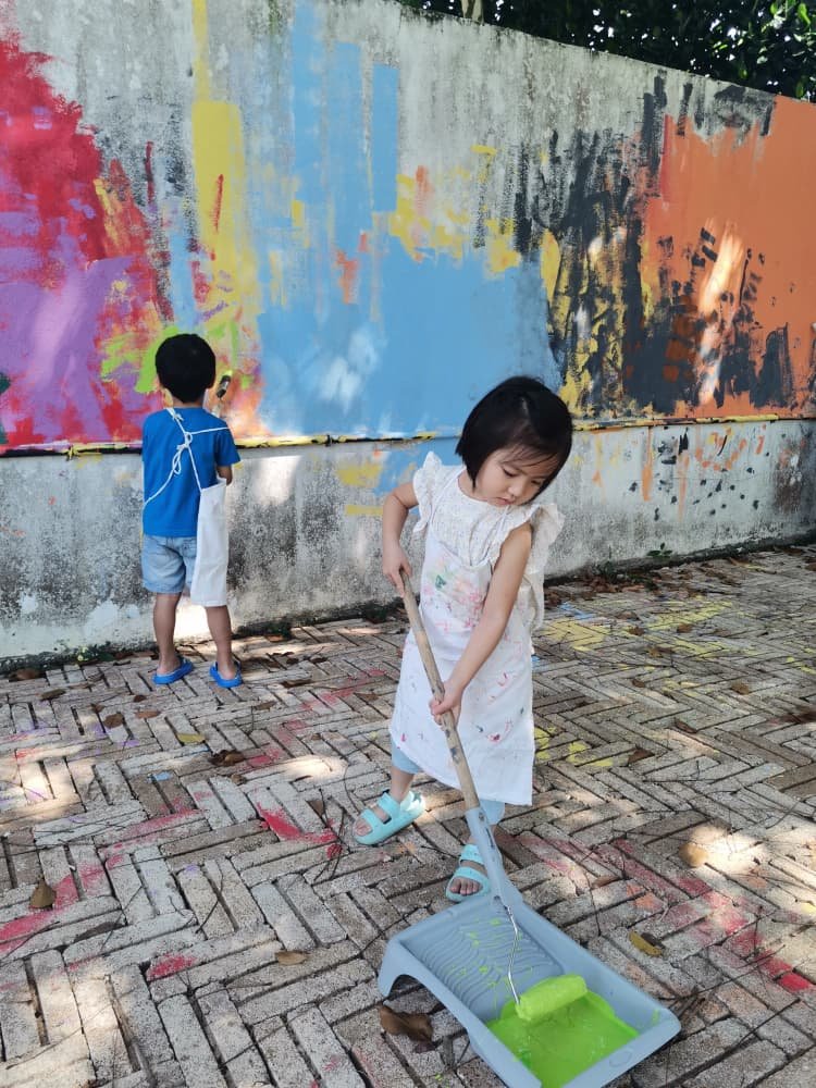 Two children painting a mural on a wall outdoors. One girl is sweeping with a dustpan filled with green liquid, and a boy is painting with a brush in the background.
