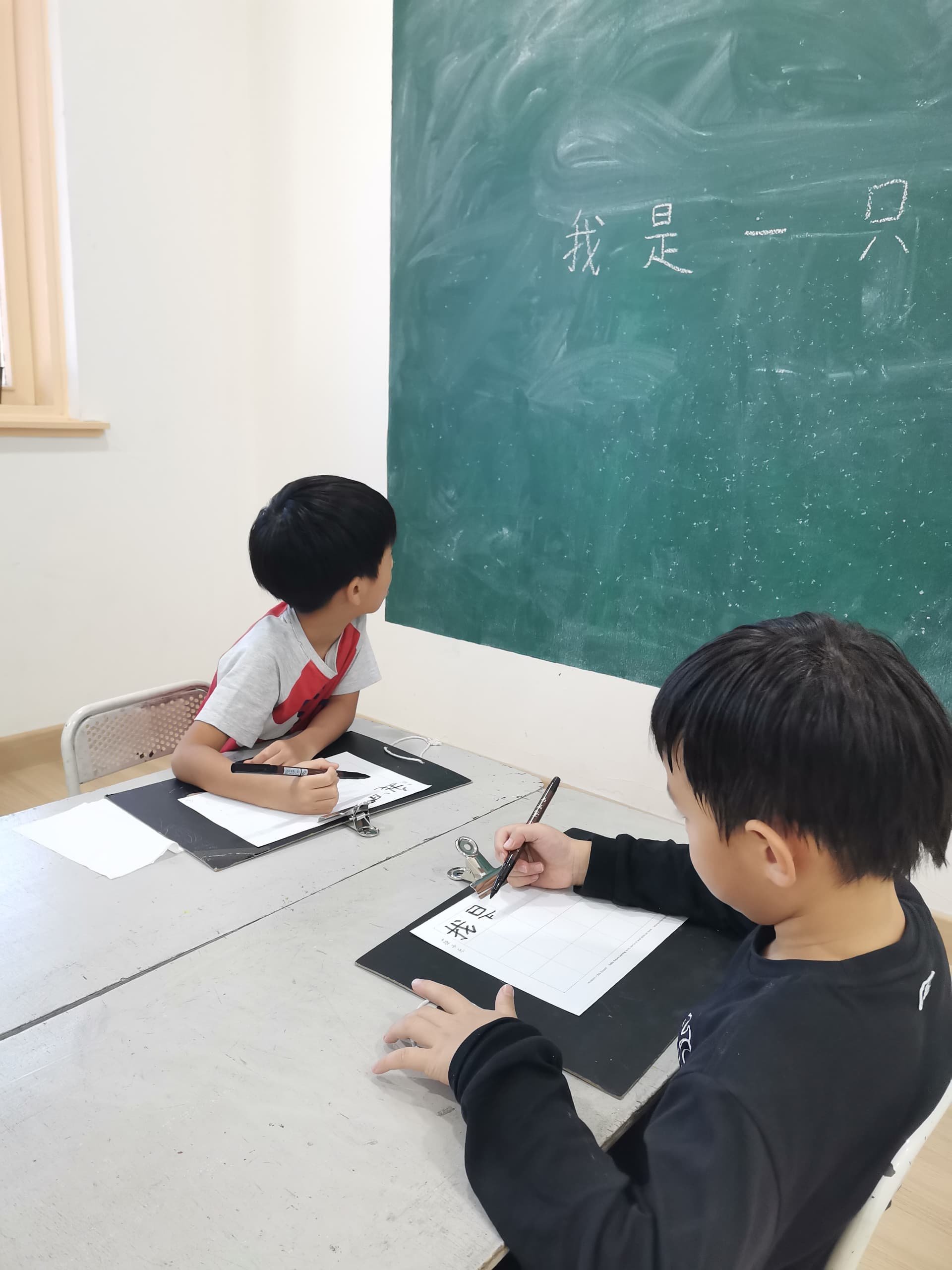 Two children sitting at a table, practicing Chinese calligraphy with black ink brushes, in front of a green chalkboard with Chinese characters written on it.