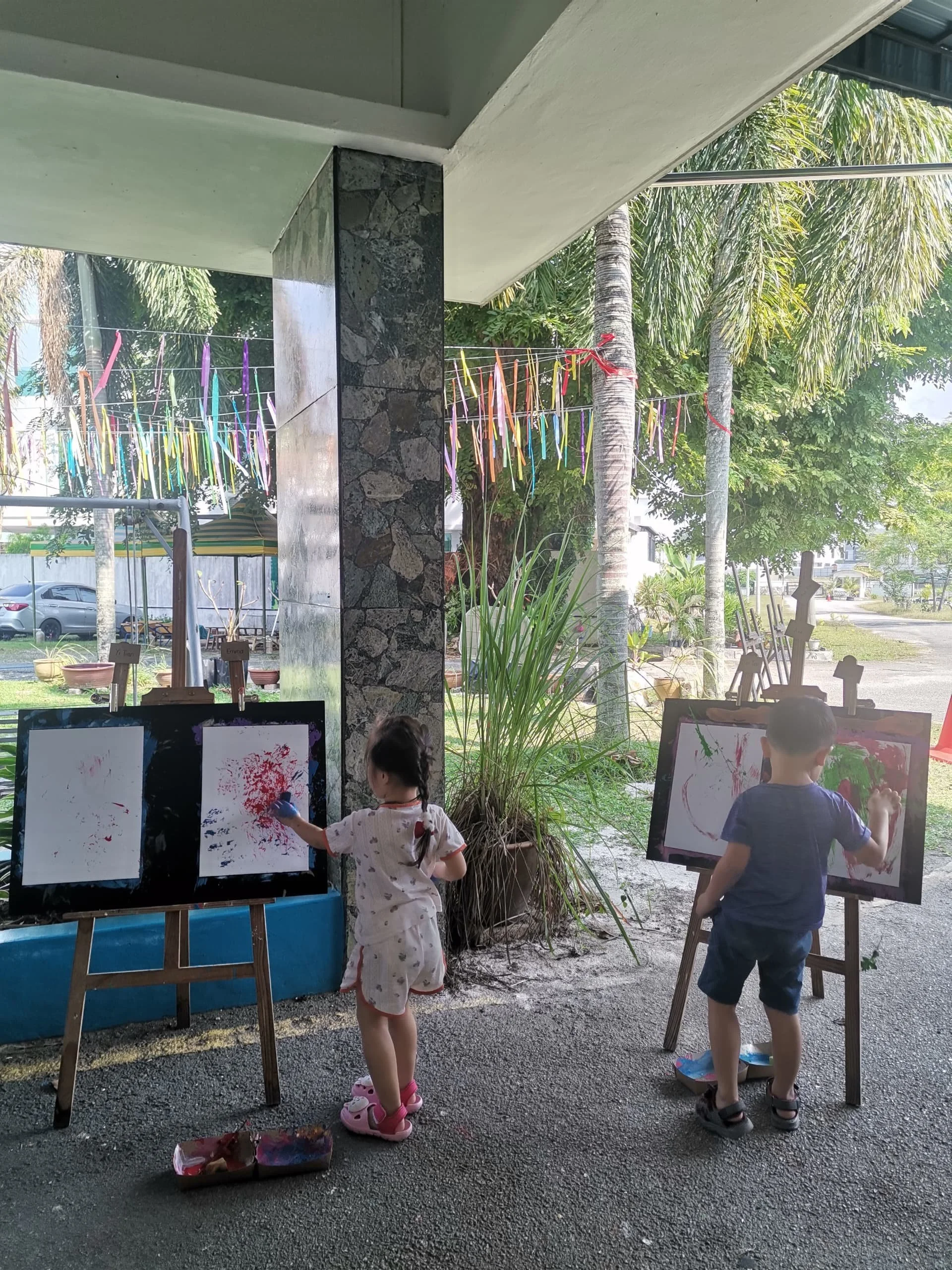 Two children painting on easels outside under a covered patio, with trees and colorful ribbons hanging in the background.