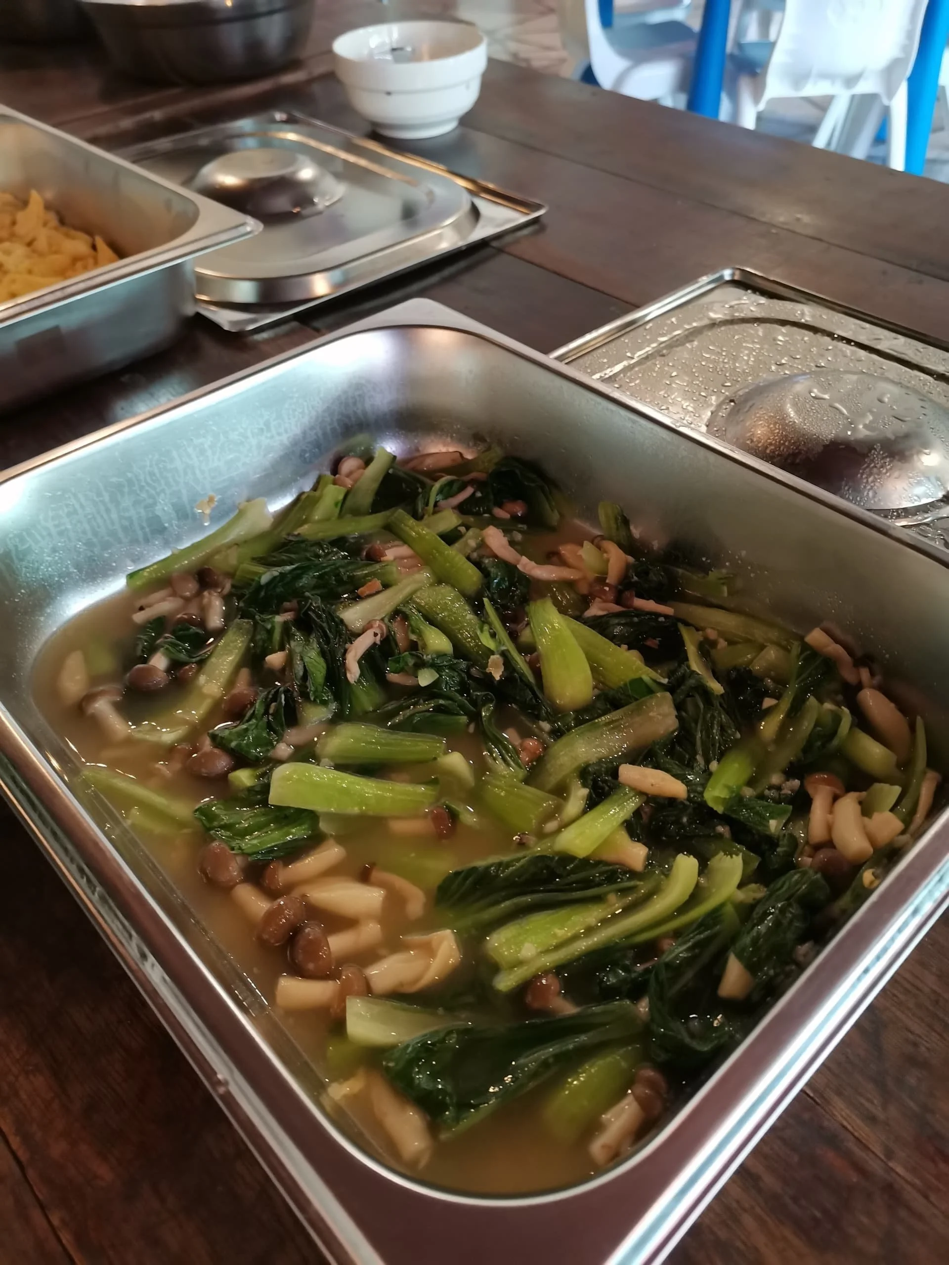 Stainless steel serving tray with mixed greens and mushrooms, placed on a dark wooden table, with other trays, bowls, and a high chair visible in the background.