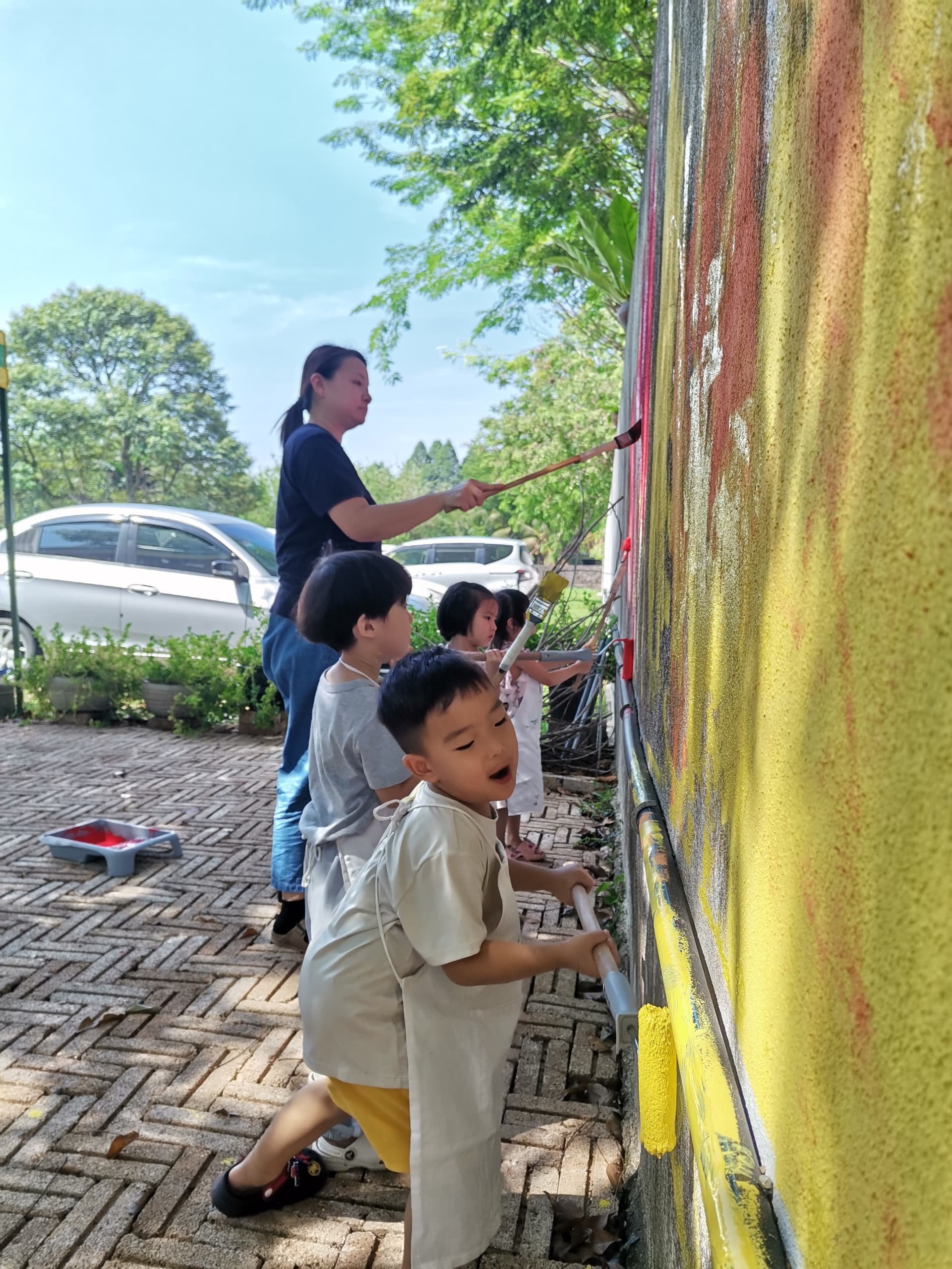 Children and a woman painting a large outdoor mural on a yellow wall with paint rollers, in a park-like setting with cars parked in the background.