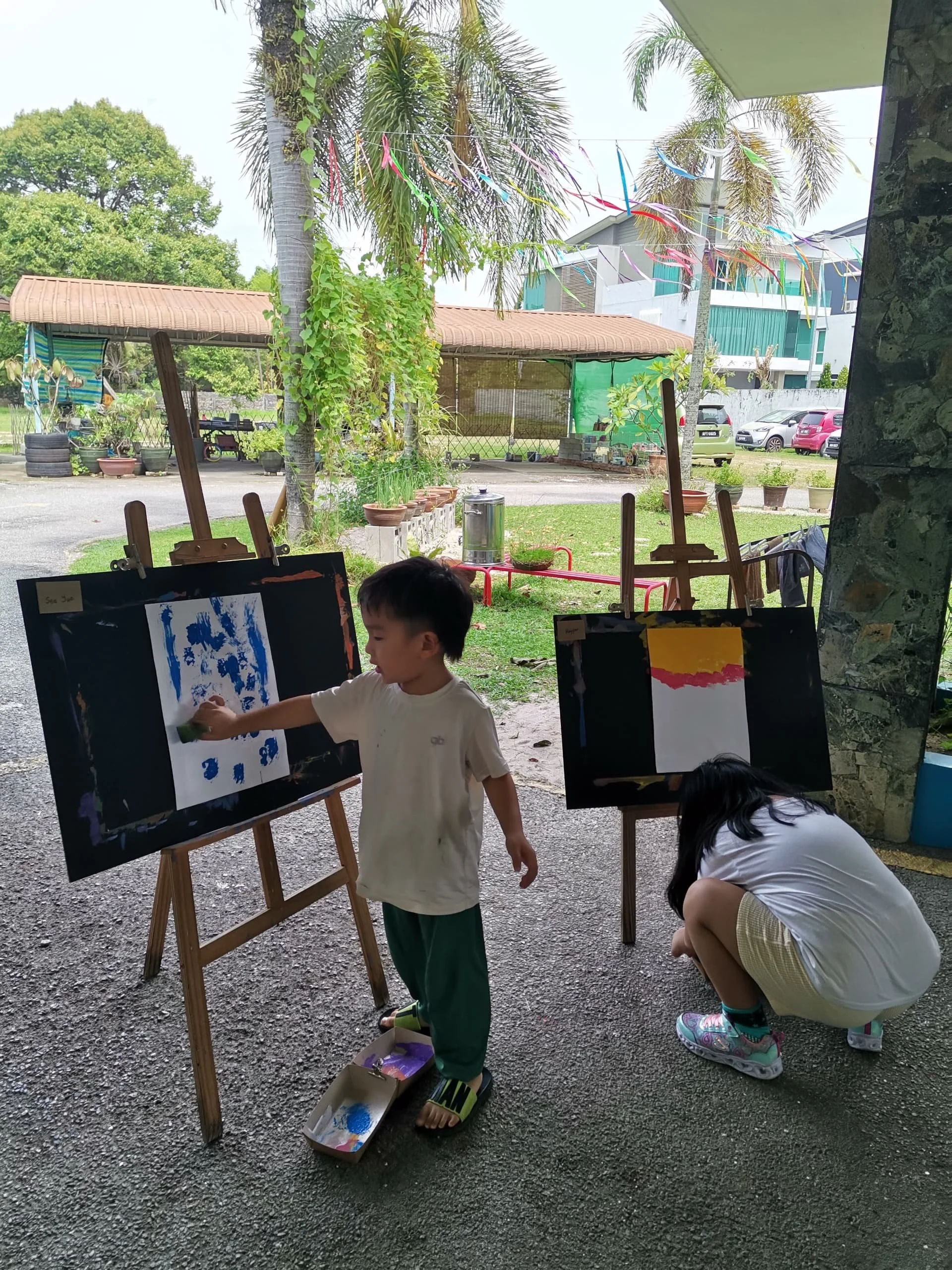 Two children are painting on easels outdoors in a garden area. The boy is standing and painting with blue and green colors; the girl is crouched down, possibly cleaning brushes or setting up art supplies. The background features trees, potted plants,