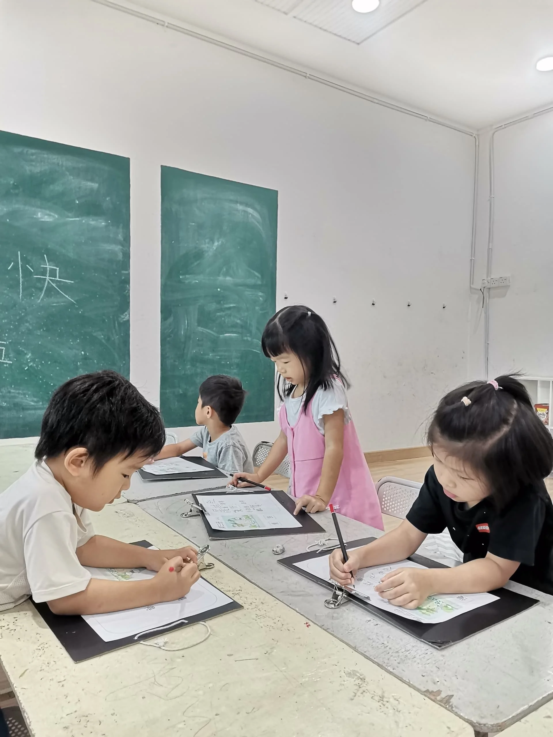 Children in a classroom sitting at a table with clipboards, writing or drawing on paper, with some standing and some sitting. There are blackboards on the wall behind them and a light-colored ceiling with fluorescent lights.