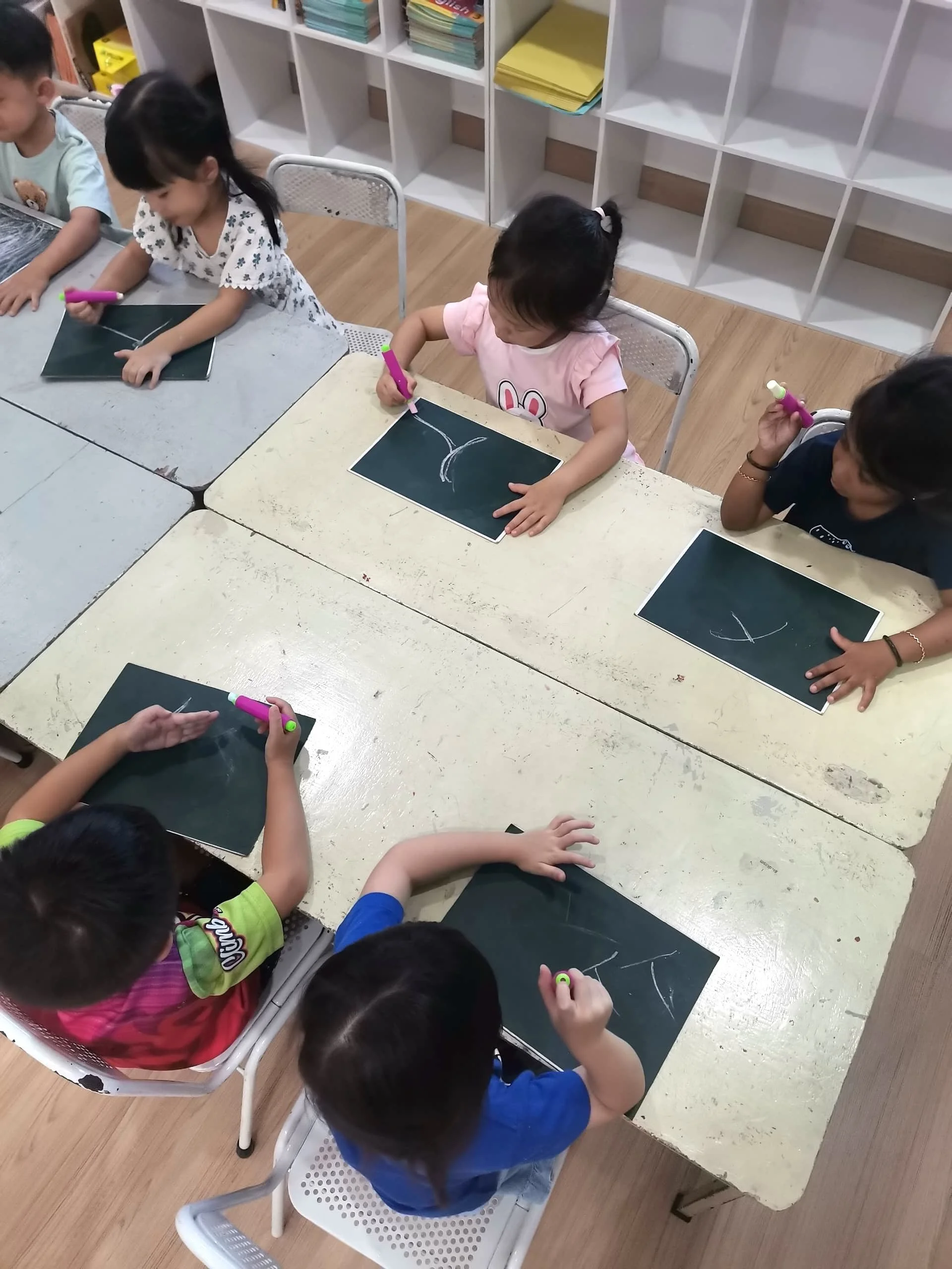 Children sitting at a table drawing on blackboards with white chalk in a classroom.