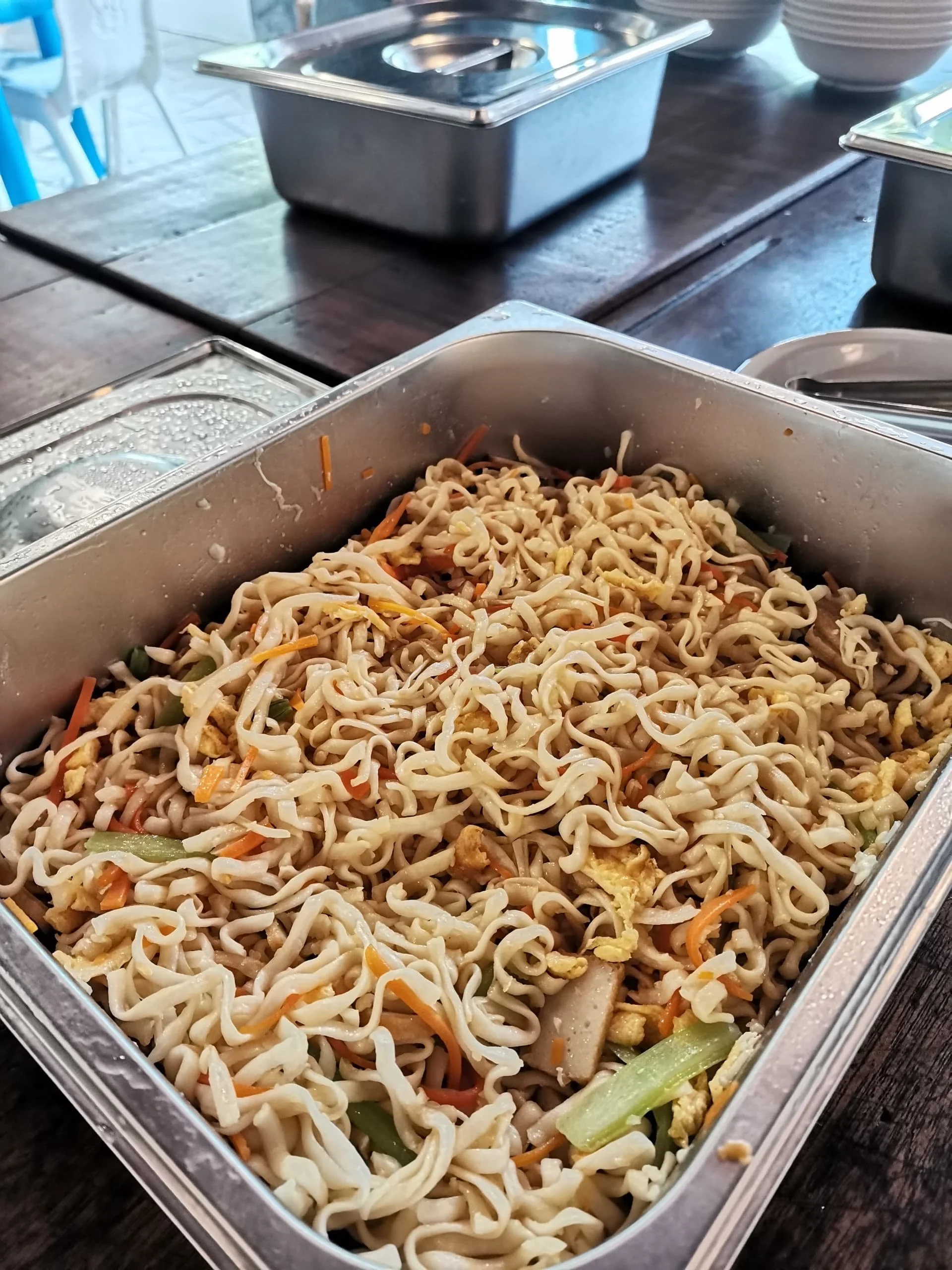 A metal container filled with cooked ramen noodles mixed with vegetables and scrambled eggs, placed on a dark wooden table.